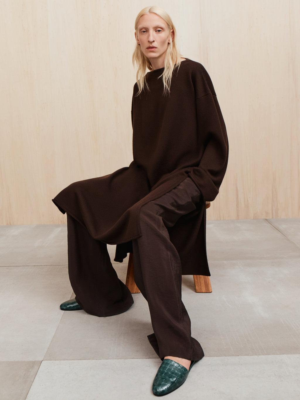 Person seated on wooden stool wearing an oversized dark brown tunic and wide brown pants with green textured loafers