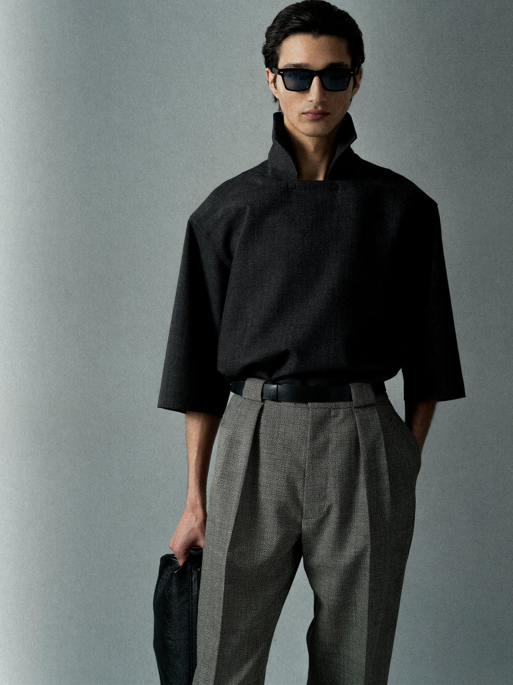 Person wearing a loose black top with a high collar and wide sleeves paired with gray pleated trousers and sunglasses