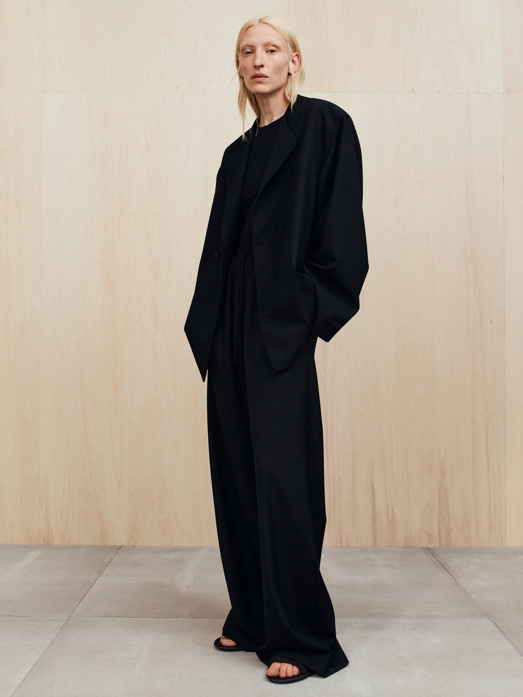 Person with pale blonde hair wearing an oversized black suit with wide-leg pants standing against a light wood wall
