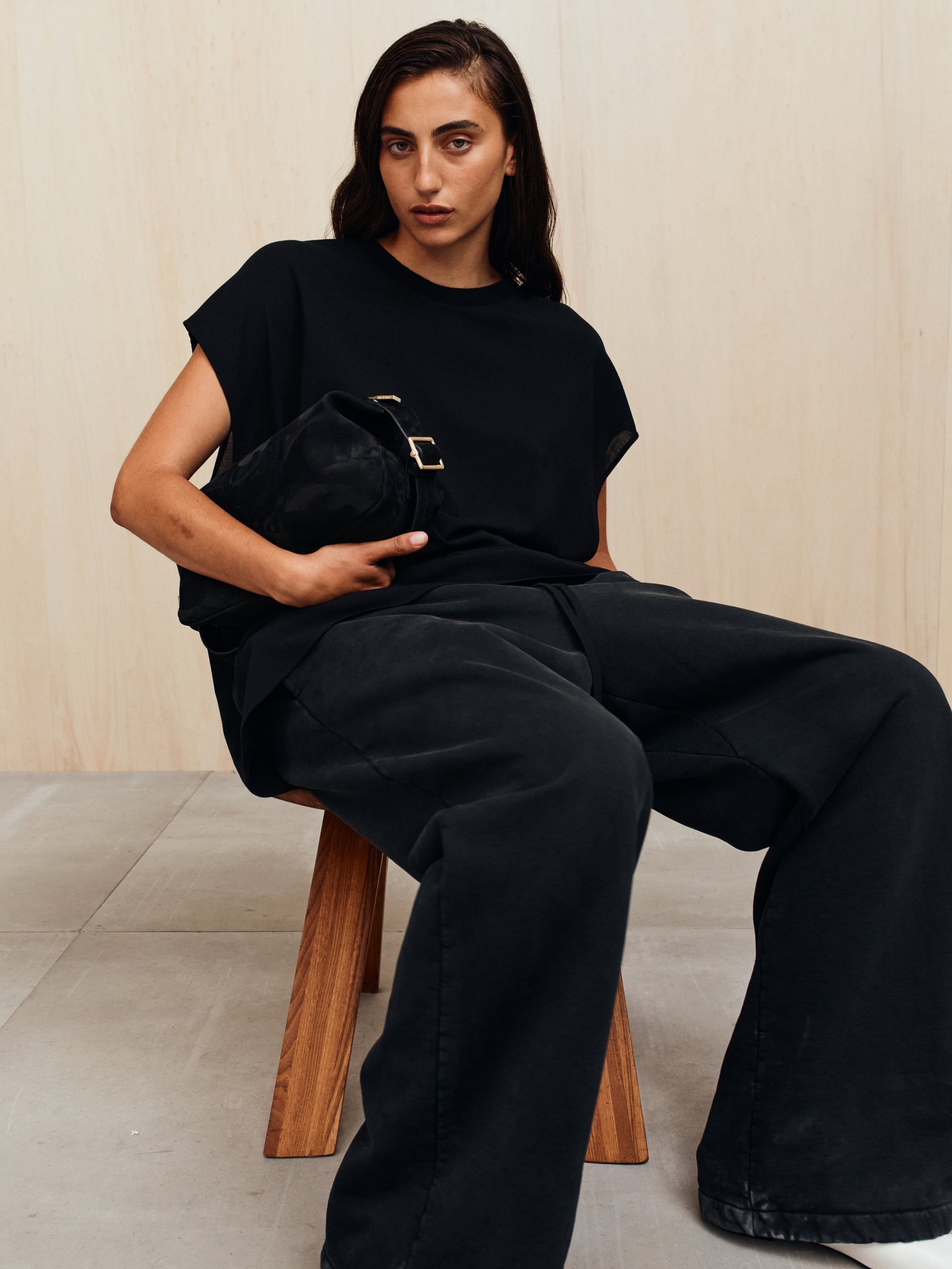 Person with medium-length dark hair wearing a loose black top and wide black pants seated on a wooden stool