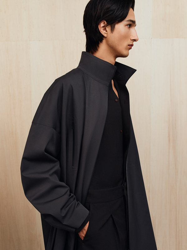 Profile of a man wearing a high-collar dark coat over a black shirt and black pants against a light wood background