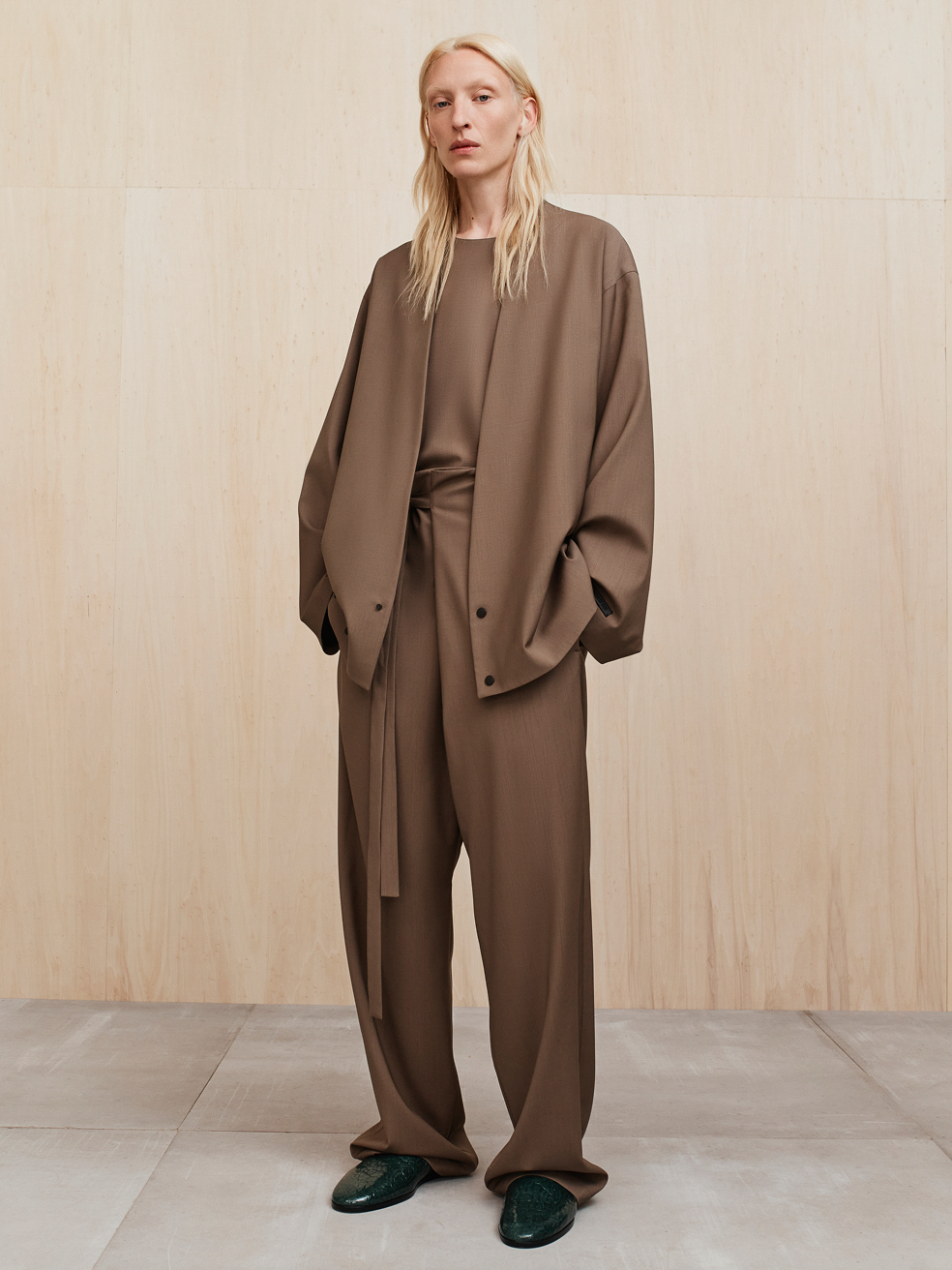 Tall person with long blonde hair wearing loose brown wide-legged pants and matching oversized jacket standing indoors
