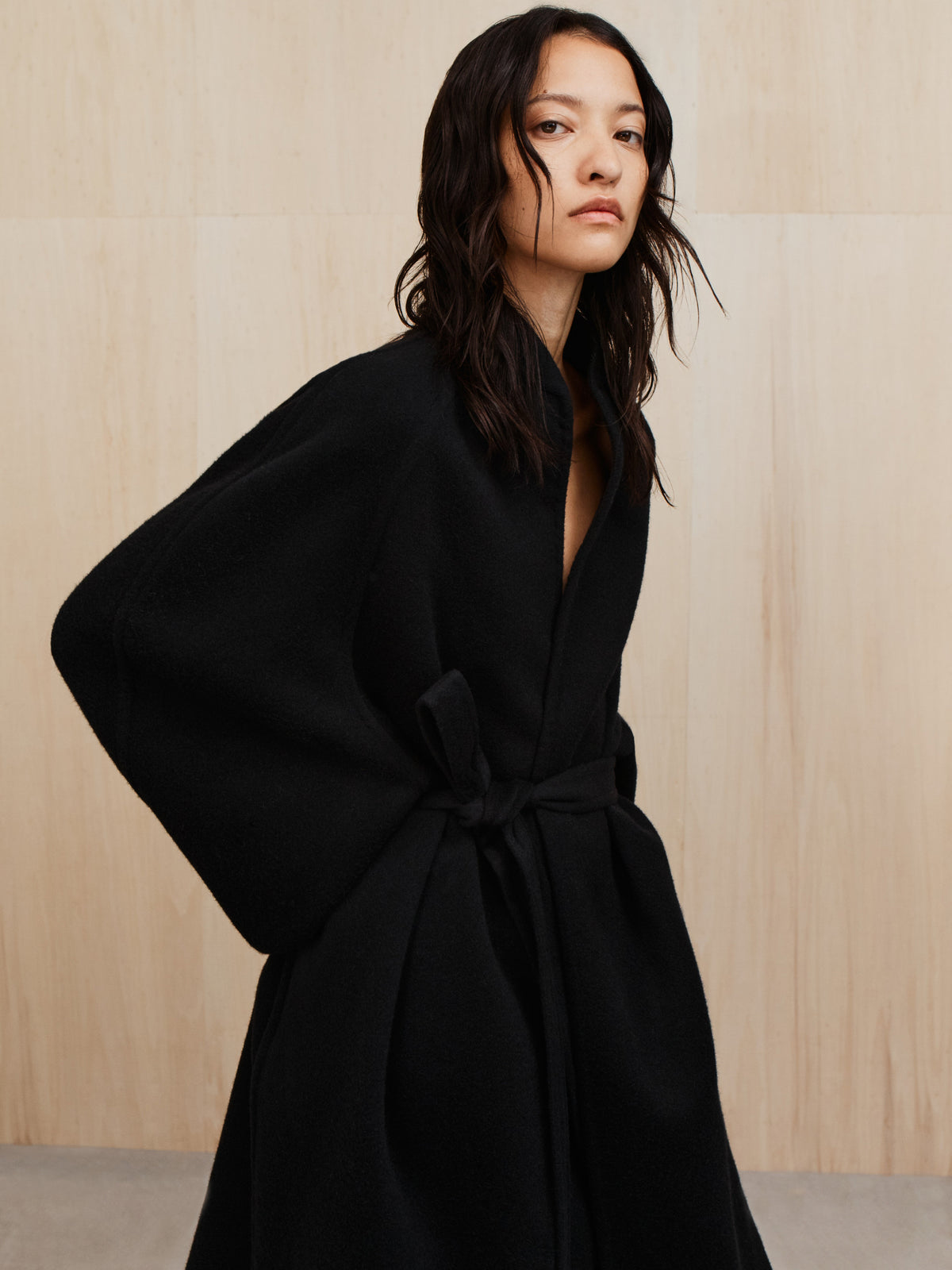 Person with long dark wavy hair wearing a black belted coat standing against a light wood panel wall
