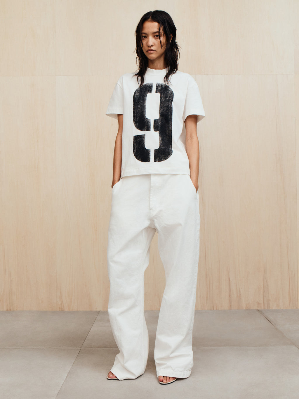 Person with dark wet-look hair wearing a white t-shirt with a large black number 9 and loose white pants standing on a tiled floor