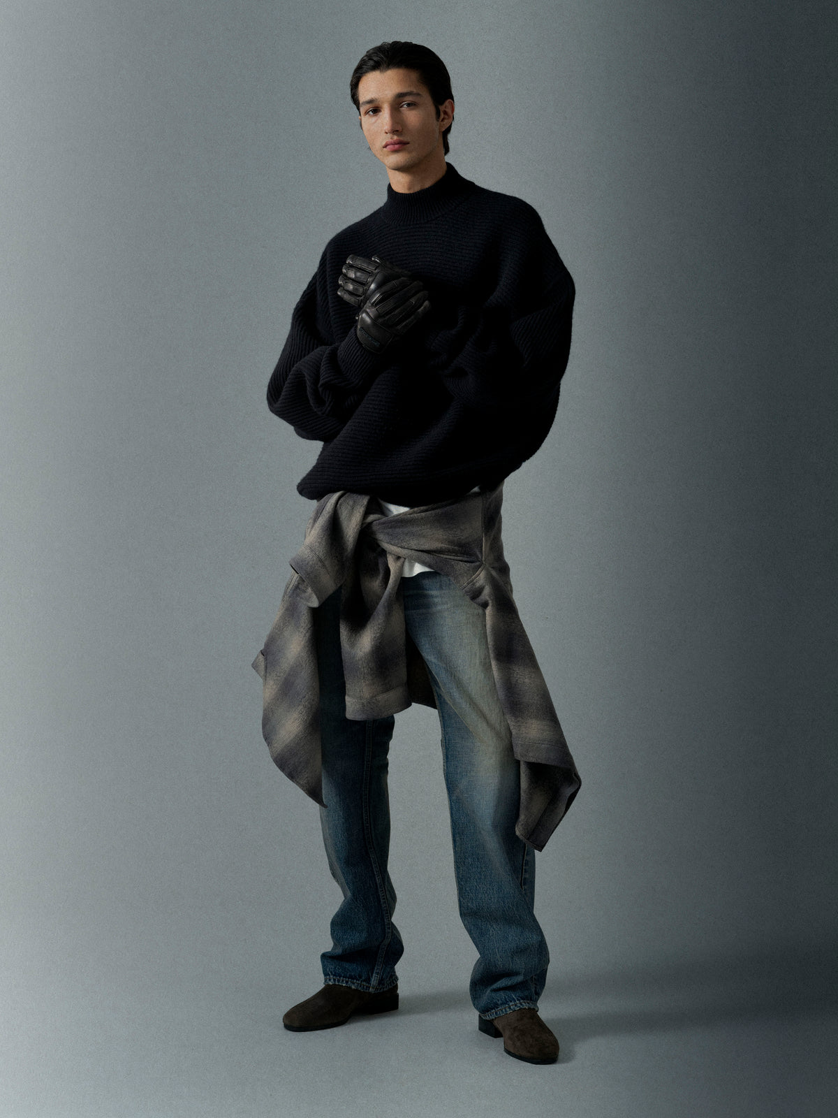 Man in a loose black sweater with leather gloves and a plaid shirt tied around the waist wearing faded blue jeans against a gray background