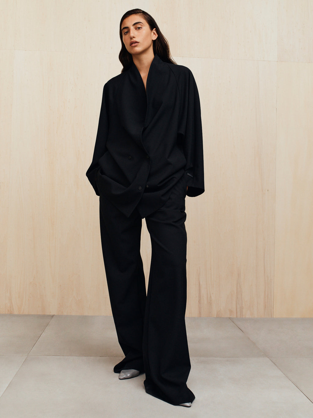Person wearing an oversized black double-breasted blazer and wide black pants standing on a pale tiled floor