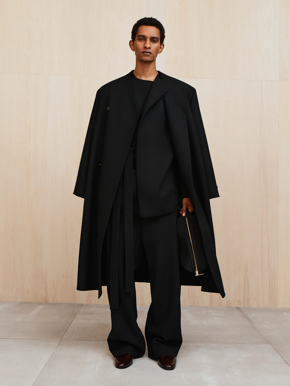 Person with short hair wearing a black long coat over a loose black suit holding a black clutch against a plain wall