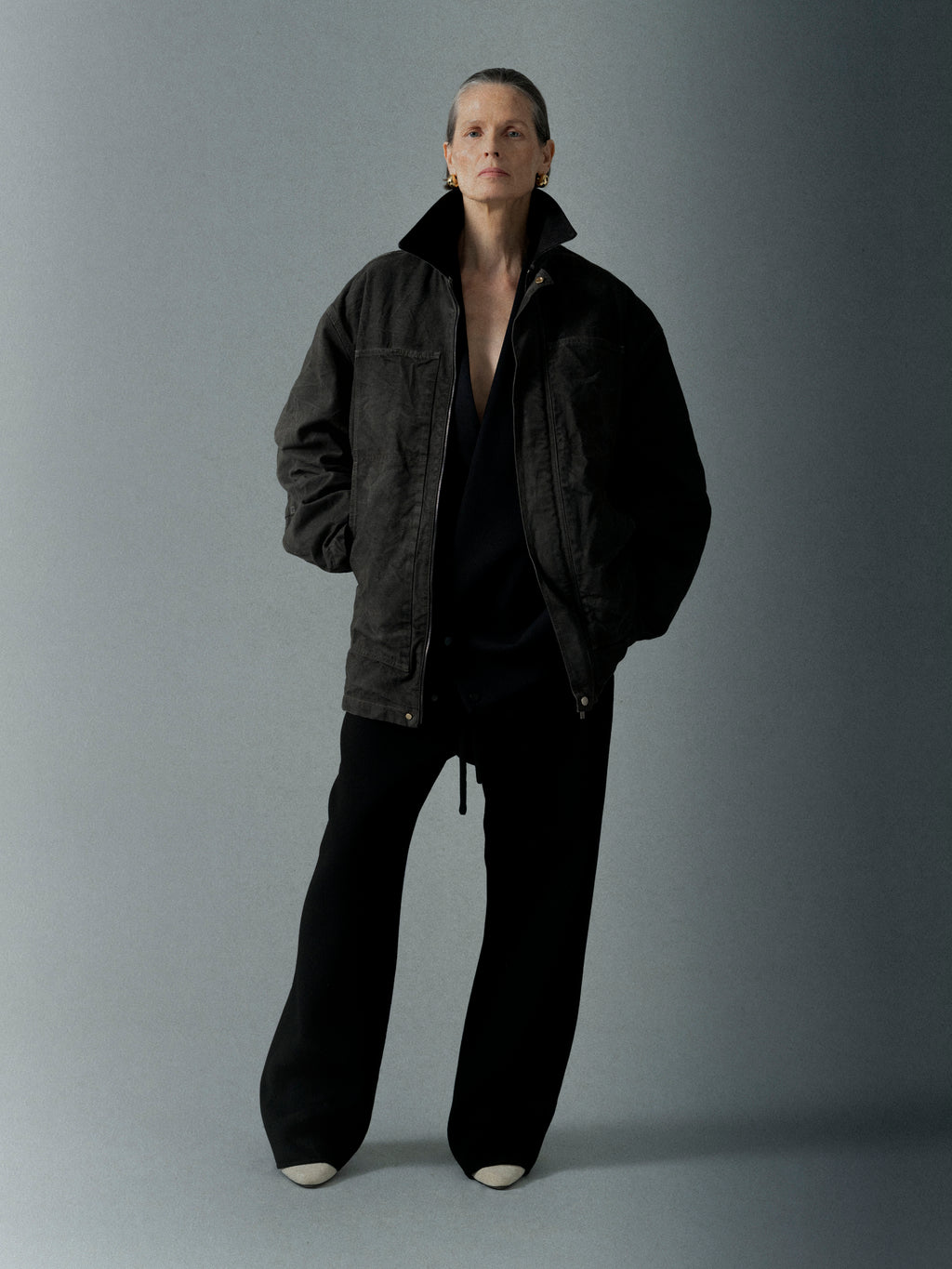 Individual wearing a dark, oversized jacket with wide black pants and light-colored shoes standing against a neutral backdrop