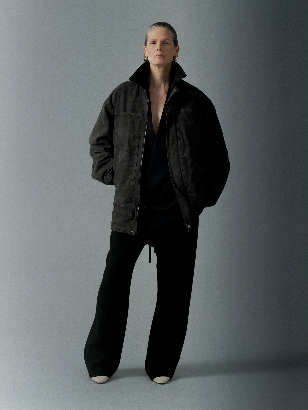 Individual wearing a dark, oversized jacket with wide black pants and light-colored shoes standing against a neutral backdrop