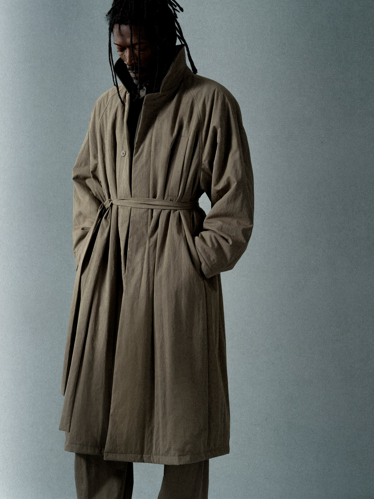 Man with dreadlocks wearing a belted oversized brown coat with high collar and hands in pockets against a plain background