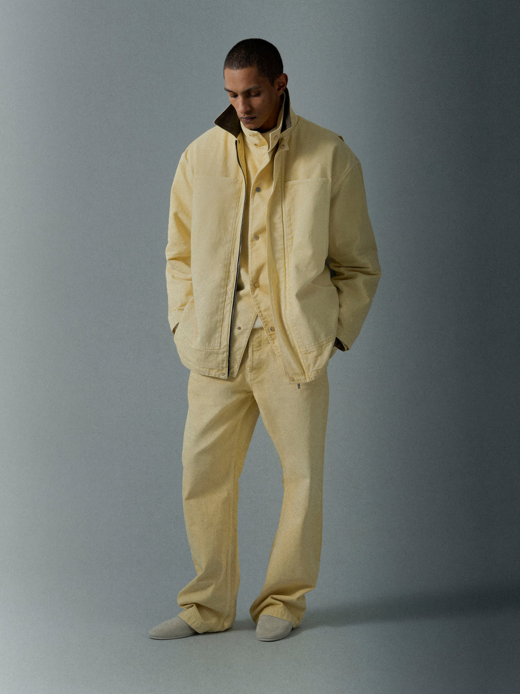 Man wearing a light beige jacket over a matching shirt and loose trousers standing against a muted gray background