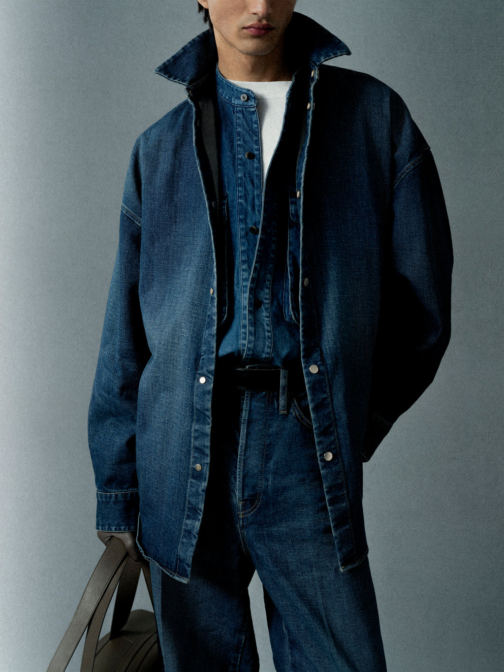 Person wearing layered dark blue denim jacket and shirt with jeans and carrying a beige bag against a plain backdrop