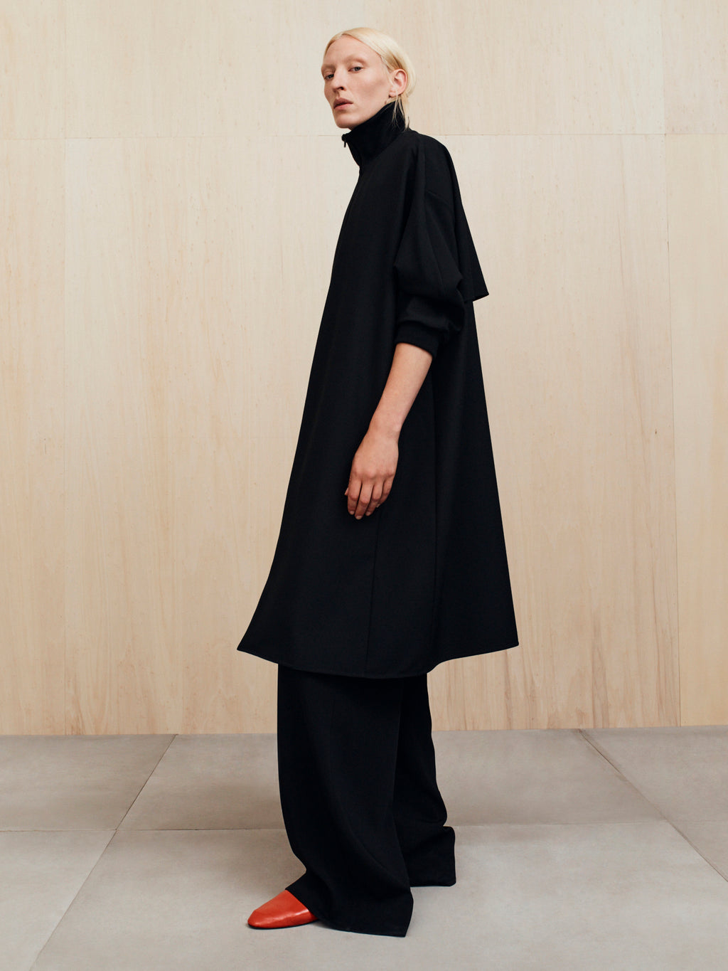 Person wearing a long loose black turtleneck tunic over wide black pants and red pointed shoes on a tiled floor