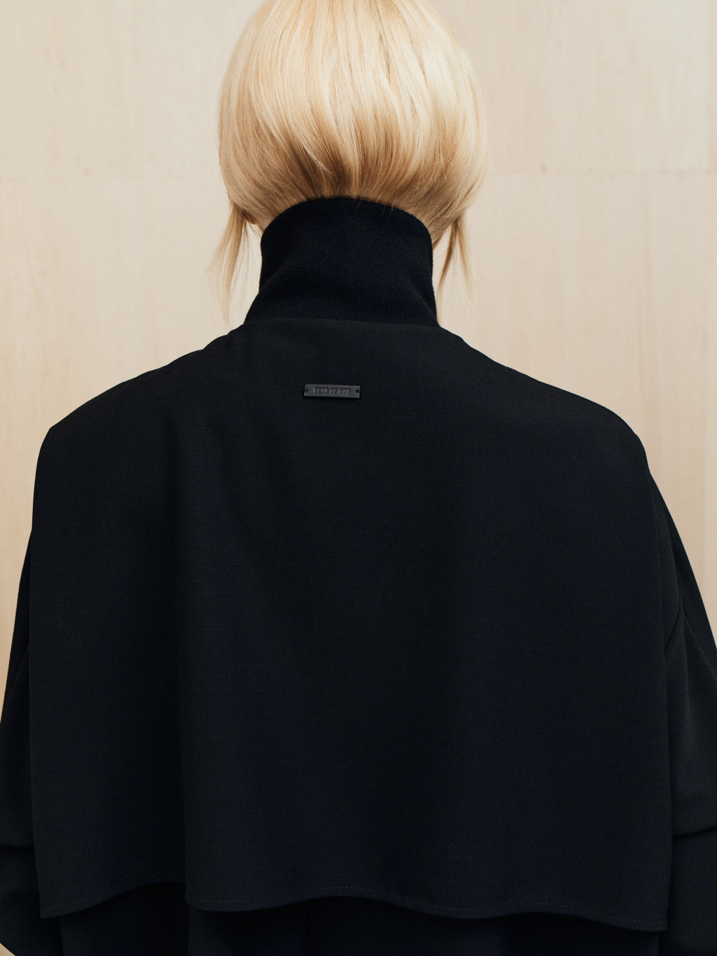 Person with short blonde hair wearing a black high-collar cape-style garment facing away from the camera
