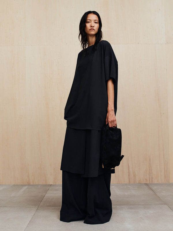 Person wearing an oversized black tunic and wide black pants holding a black bag against a wooden wall
