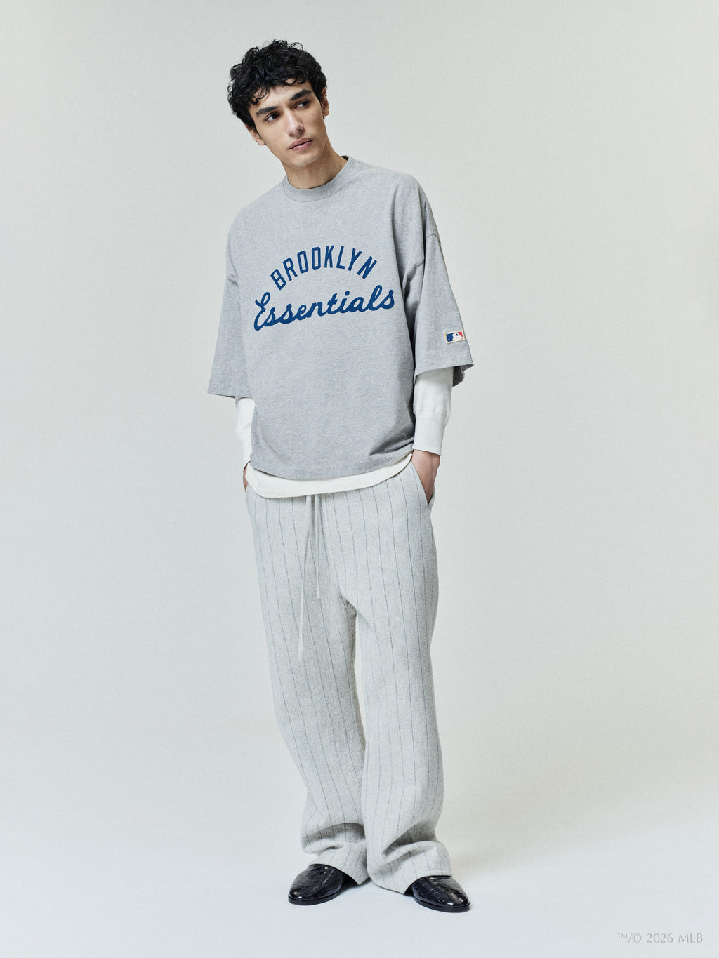Man wearing loose gray short-sleeve shirt with "Brooklyn Essentials" text and light gray pinstripe pants on neutral background