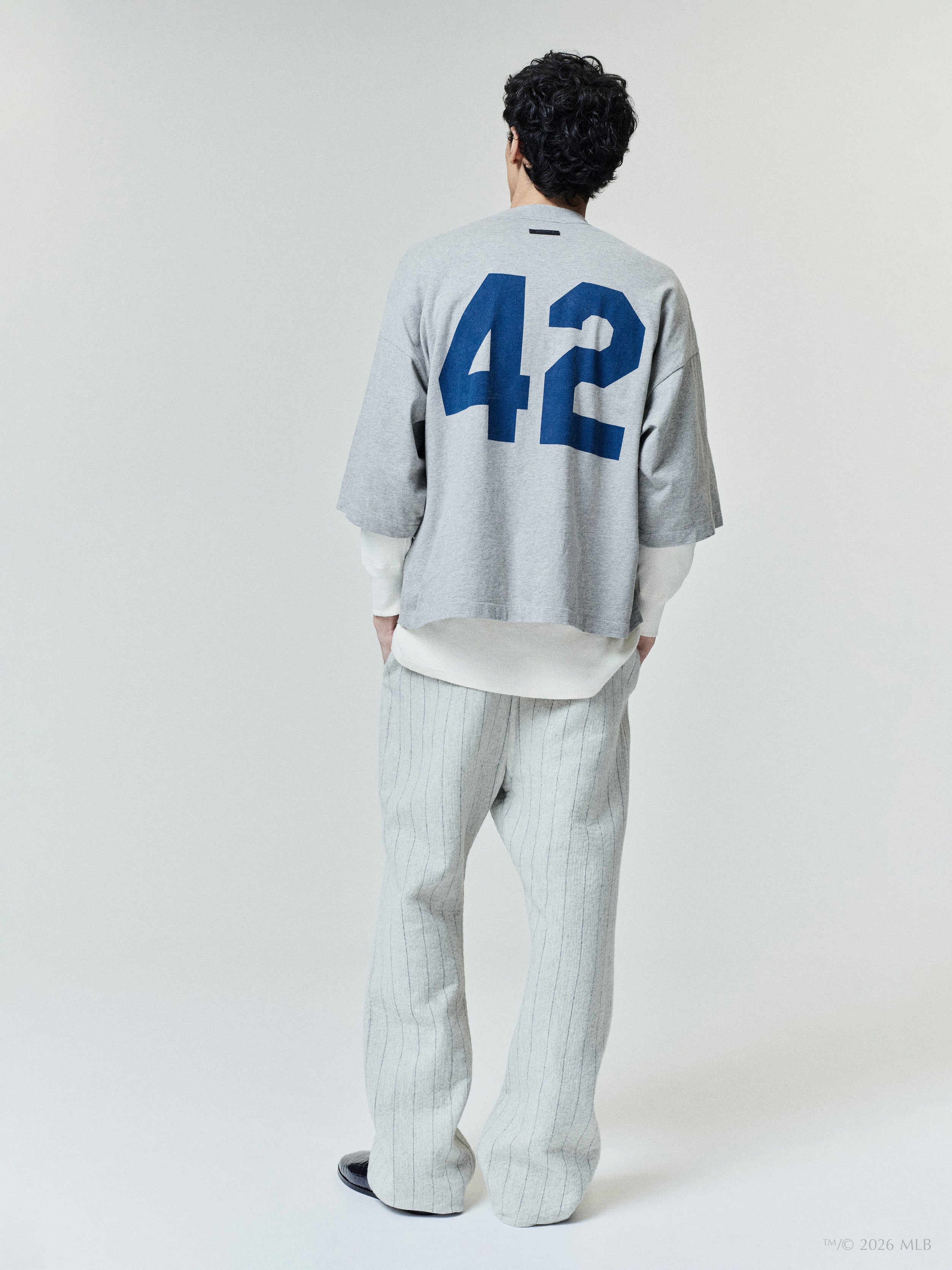 Man wearing a loose gray top with large blue number "42" on the back and light gray pinstripe pants on white background