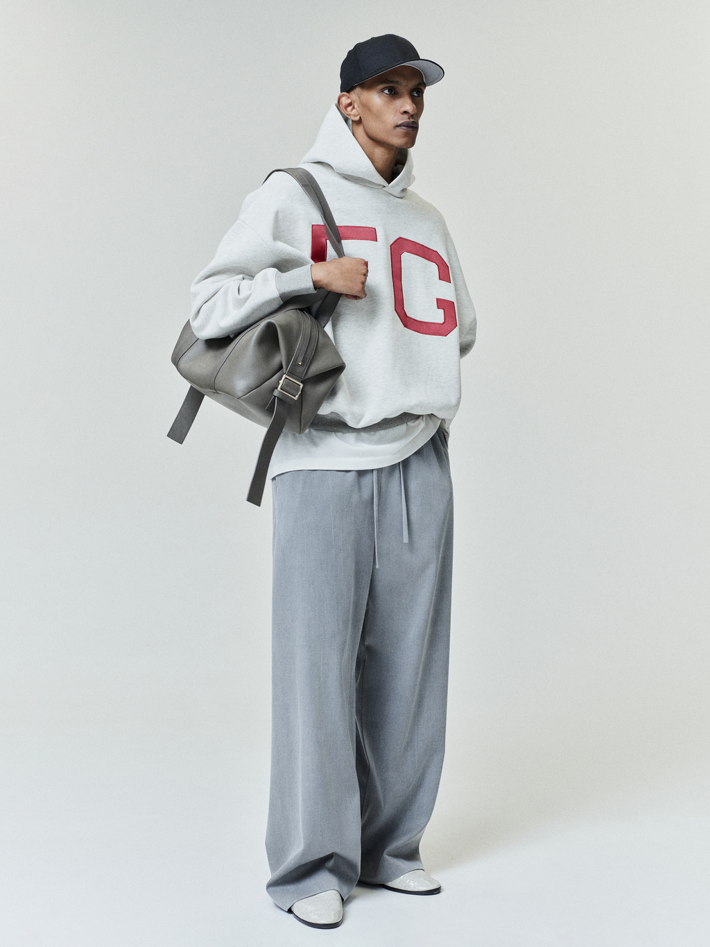 Man in light gray hoodie with bold red lettering paired with loose gray pants, white shoes, black cap, and gray backpack