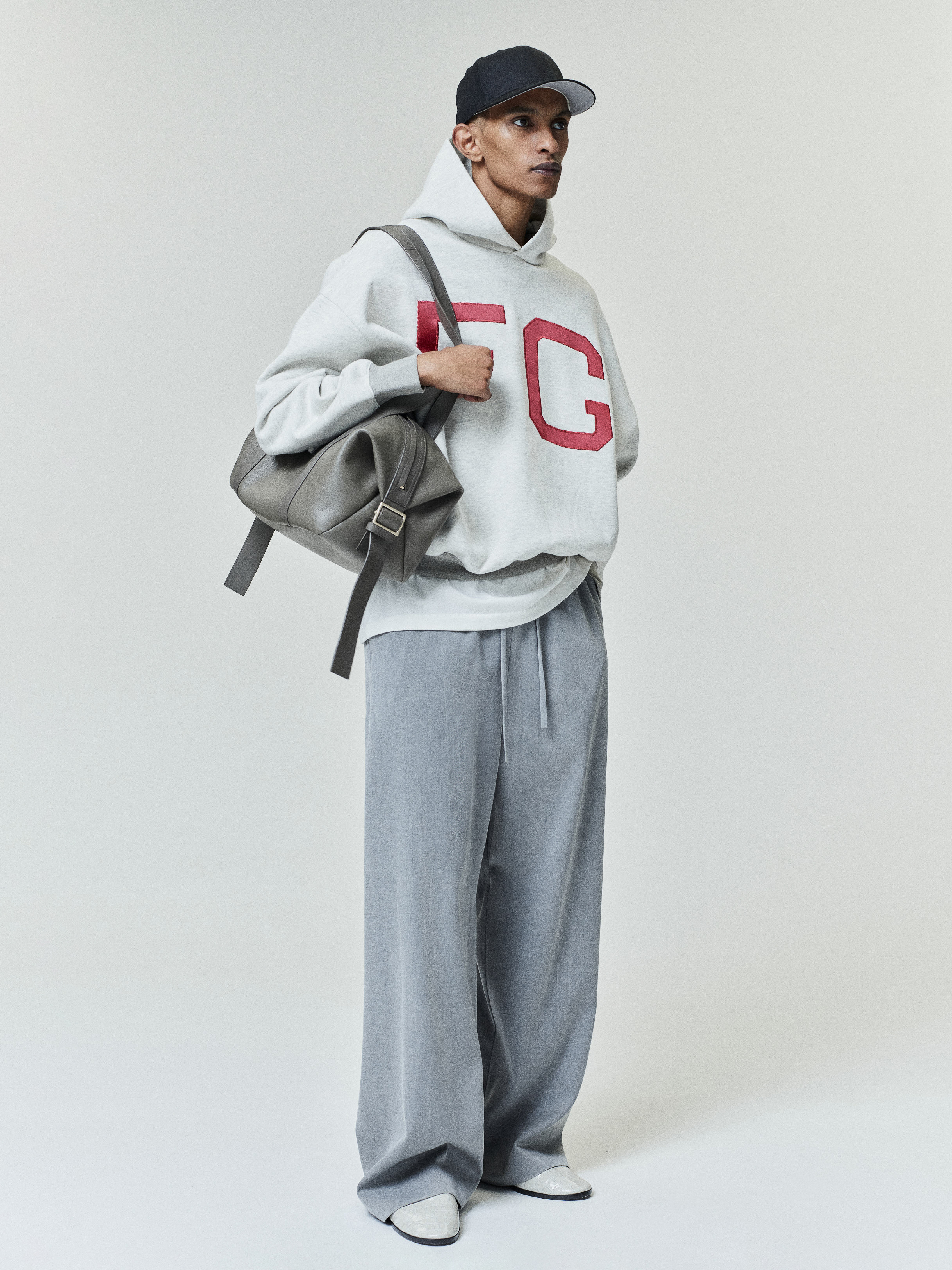 Man in light gray hoodie with bold red lettering paired with loose gray pants, white shoes, black cap, and gray backpack