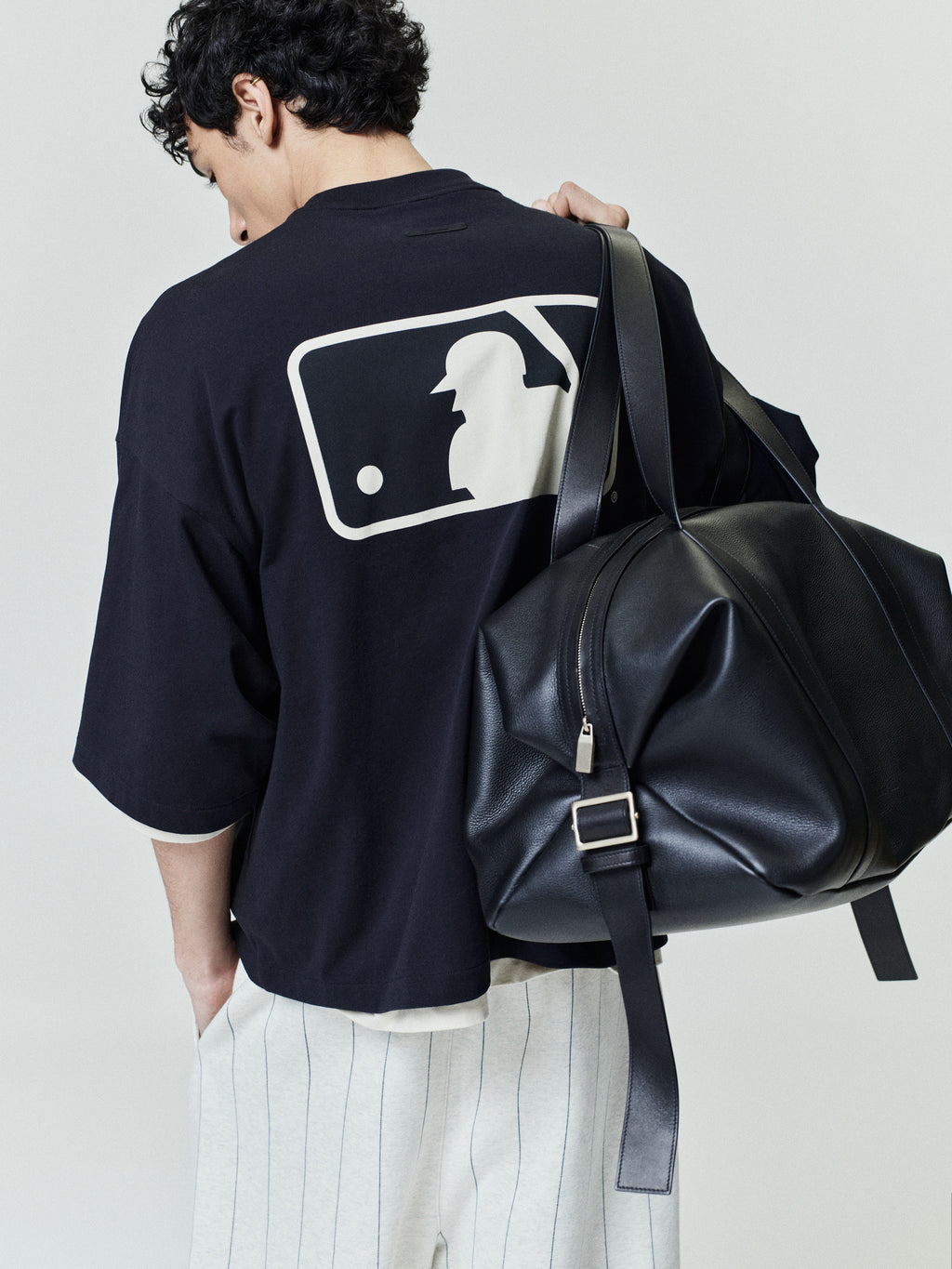 Model wearing a black MLB Merch Tee with a large white silhouette graphic of a baseball batter on the back, paired with white pinstriped pants and carrying a black leather duffel bag.