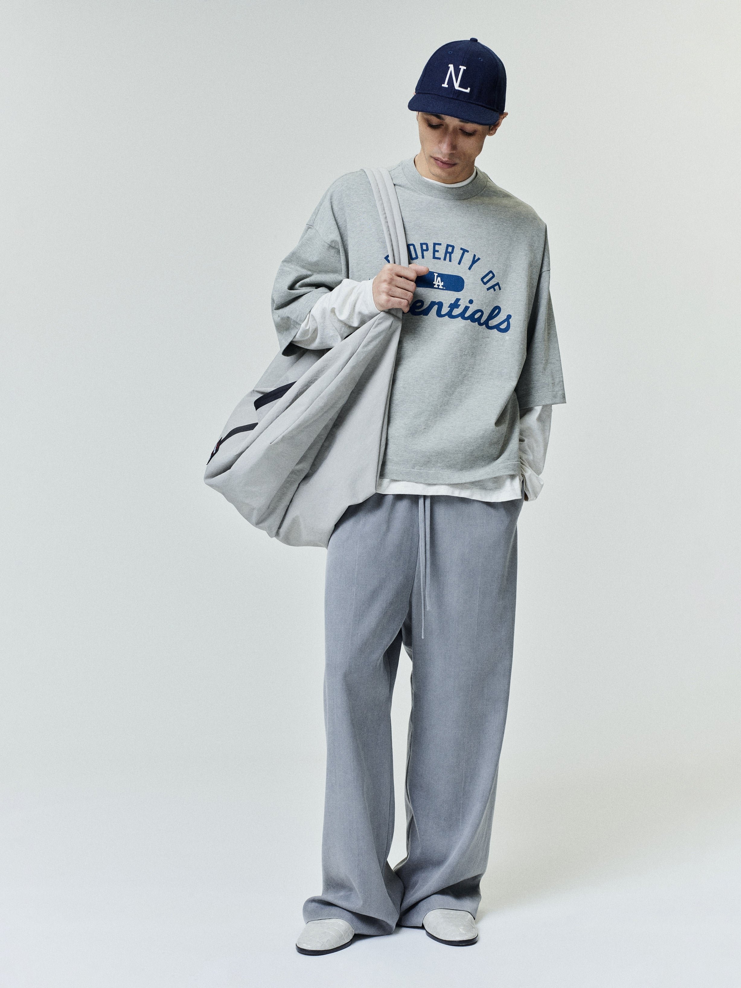 Model wearing a Dodgers Merch Tee in light gray layered over a long-sleeve white shirt, paired with loose gray pants, white sneakers, a navy cap, and carrying a large gray tote bag.