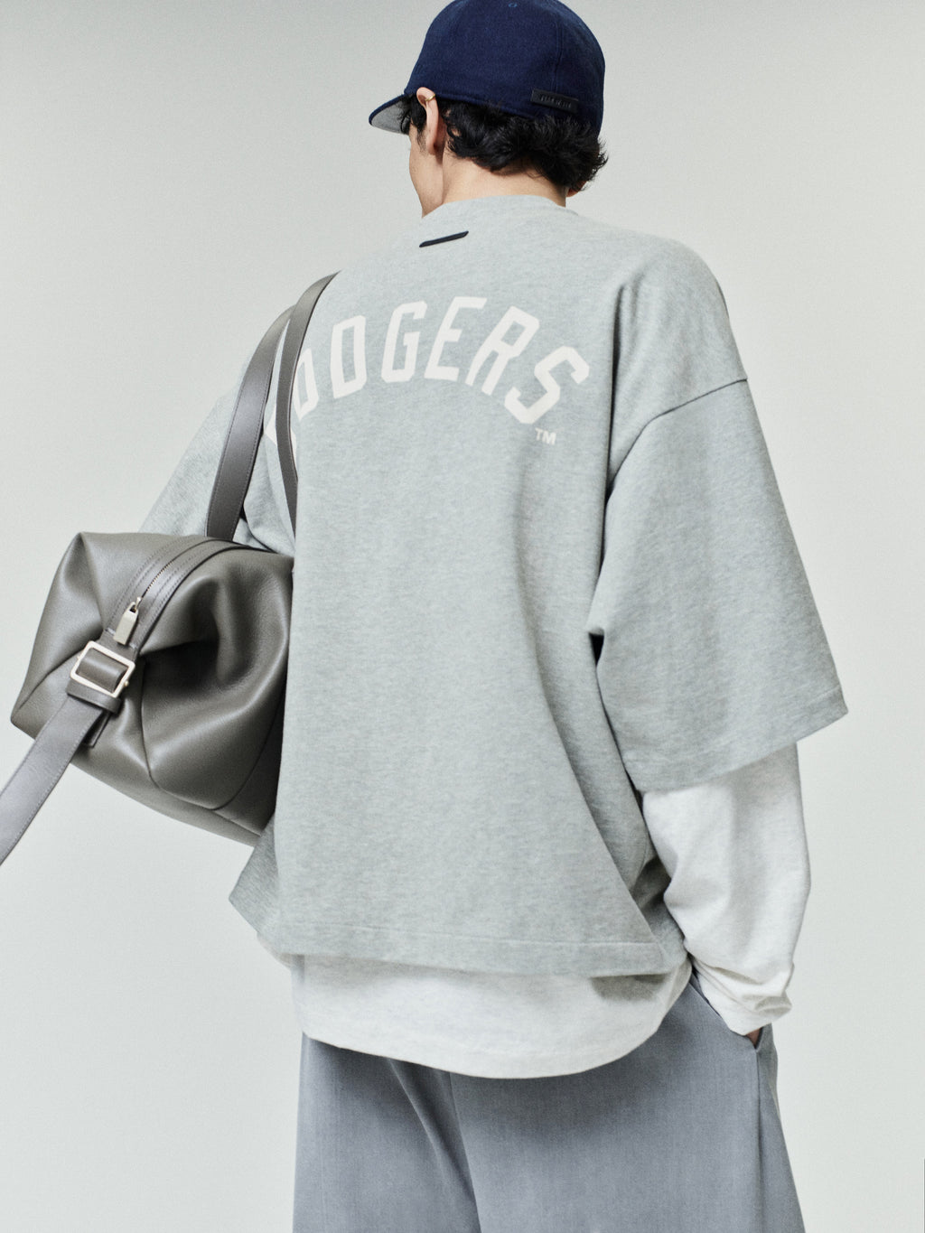 Person wearing a loose-fitting gray Dodgers Merch Tee with bold white lettering across the back, paired with layered light gray long sleeves and carrying a large gray leather duffel bag.
