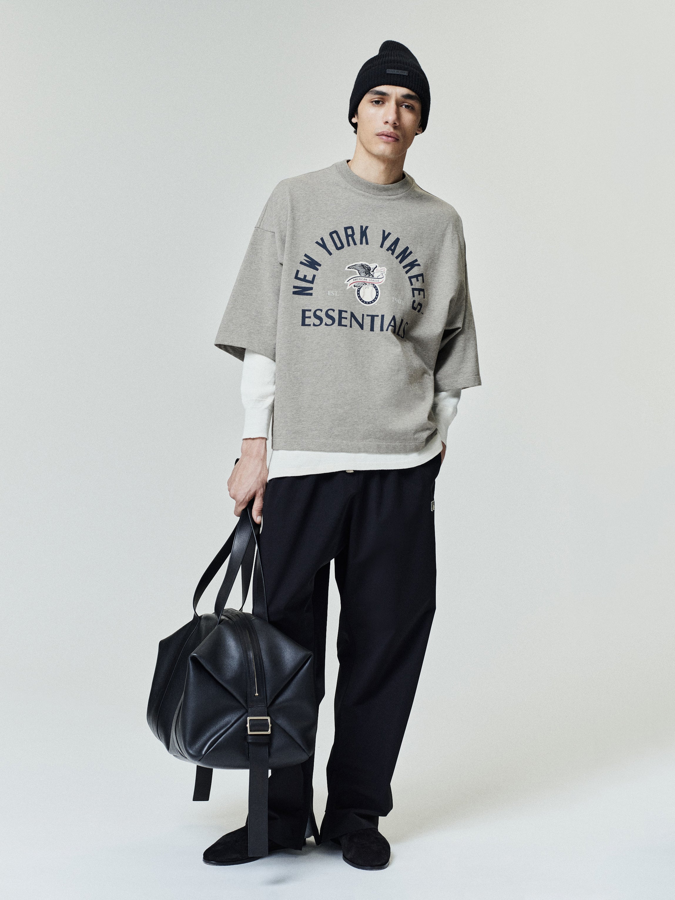 Yankees AL Merch Tee in a relaxed fit with muted gray fabric featuring navy "New York Yankees Essentials" text and graphic on a soft casual top styled with black pants and a beanie.