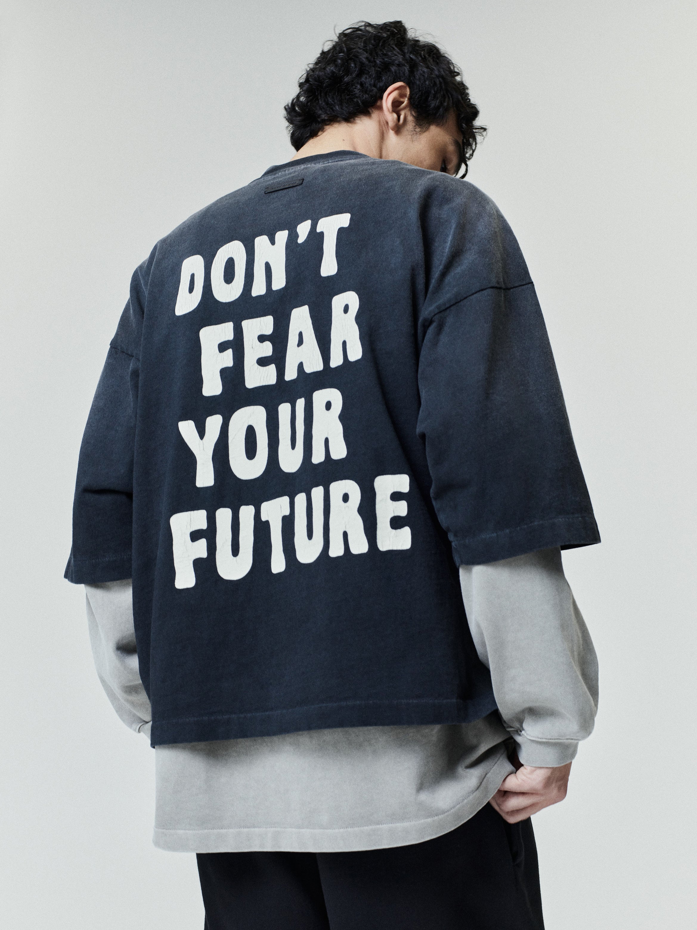 Man wearing a faded dark oversized t-shirt with bold white lettering on the back that reads "DON'T FEAR YOUR FUTURE," layered over a light gray long-sleeve shirt and paired with black pants.