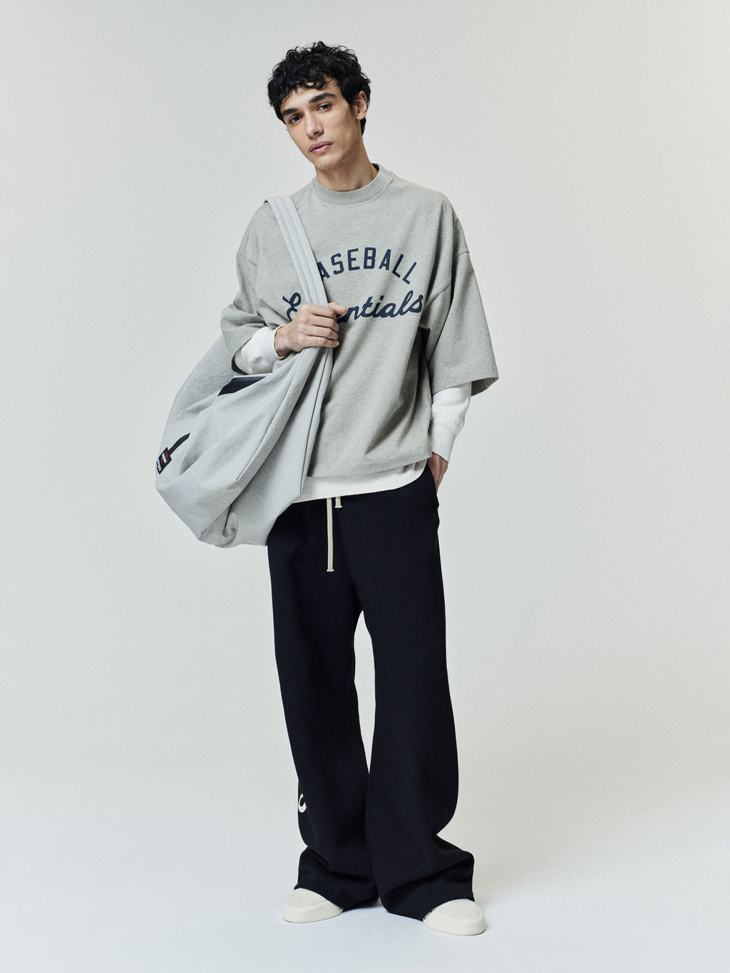 Model wearing a relaxed-fit Mlb Merch Tee in light gray with bold navy lettering, layered over a white long sleeve top, paired with black wide-leg pants and white sneakers.