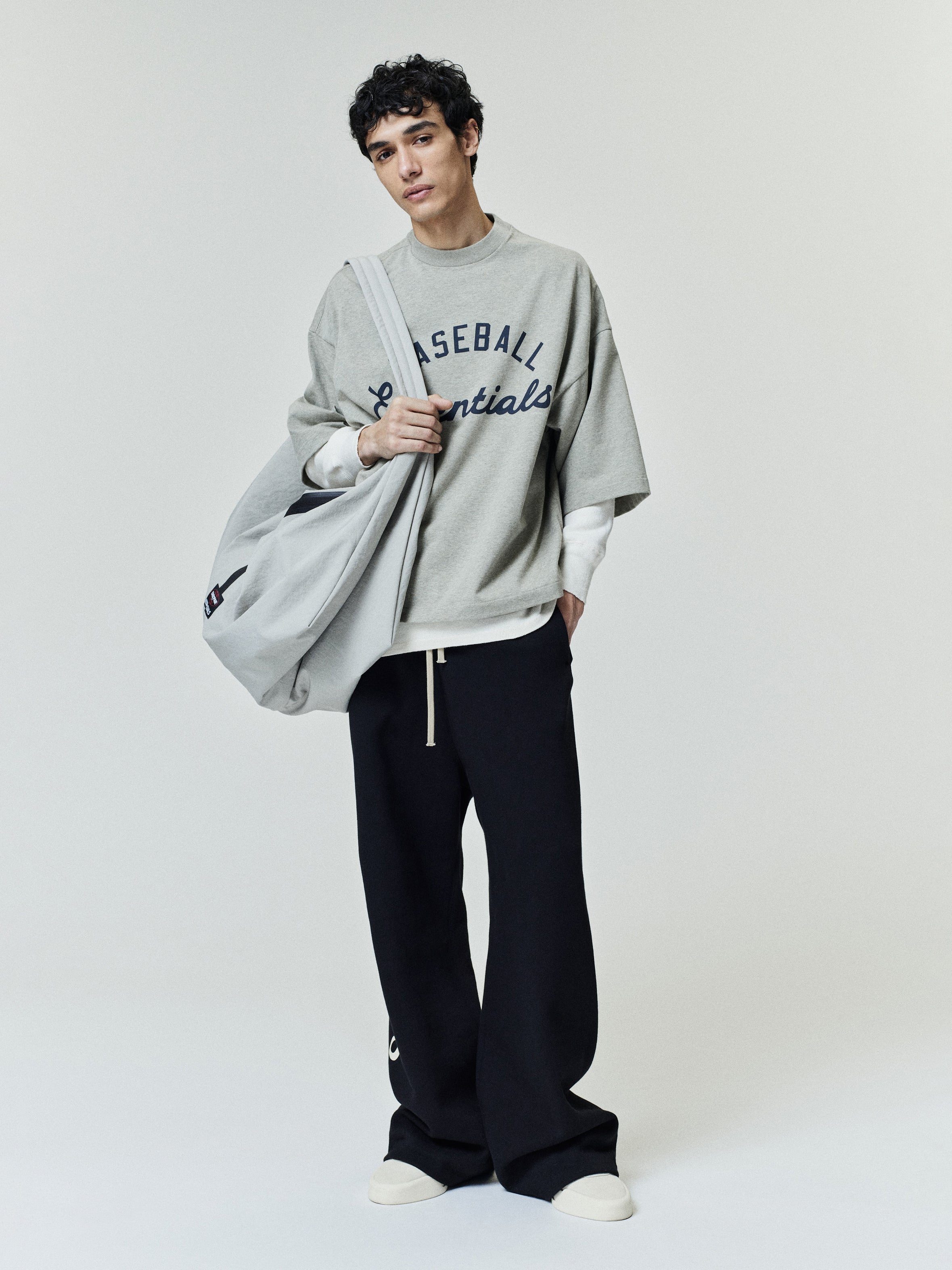 Model wearing a relaxed-fit Mlb Merch Tee in light gray with bold navy lettering, layered over a white long sleeve top, paired with black wide-leg pants and white sneakers.