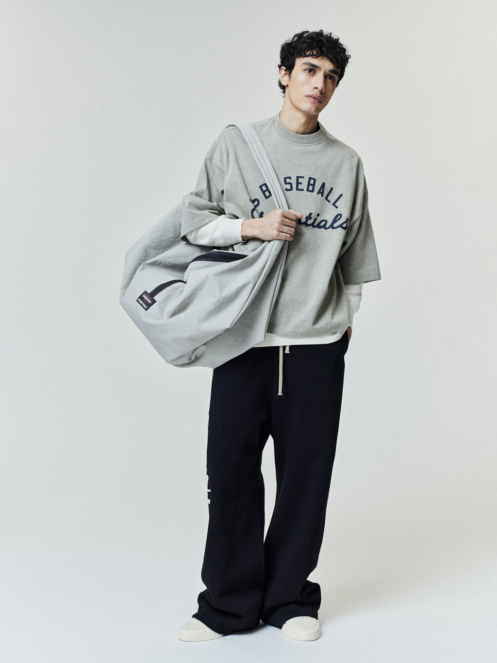 Model wearing an oversized Mlb Merch Tee in light gray layered over a white long-sleeve shirt, styled with wide-leg black pants and carrying a large gray tote bag.