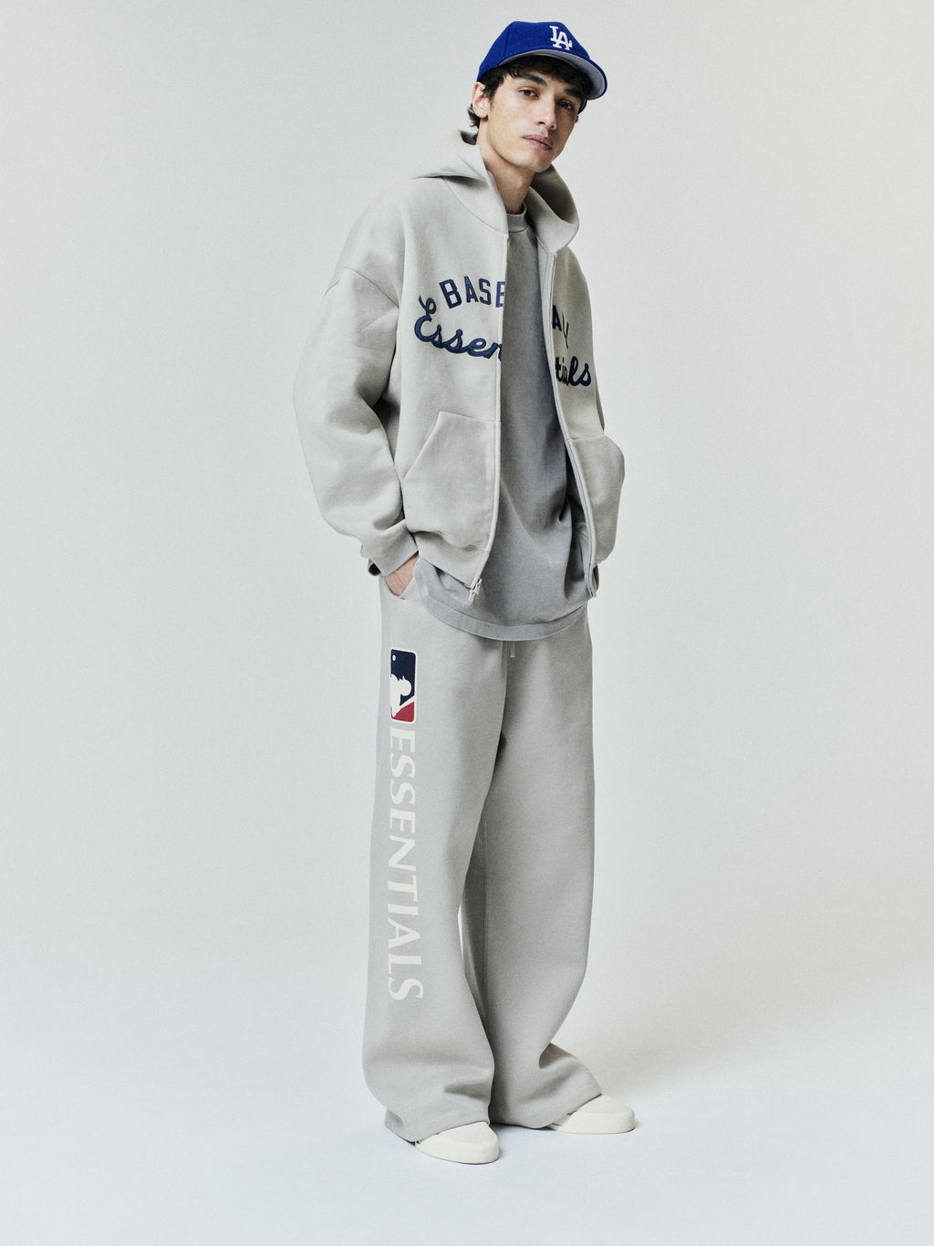 Model wearing Mlb Lounge Sweatpant in light gray with a relaxed fit and bold "ESSENTIALS" text and MLB logo on the side, paired with coordinating casual layered outerwear and sneakers.