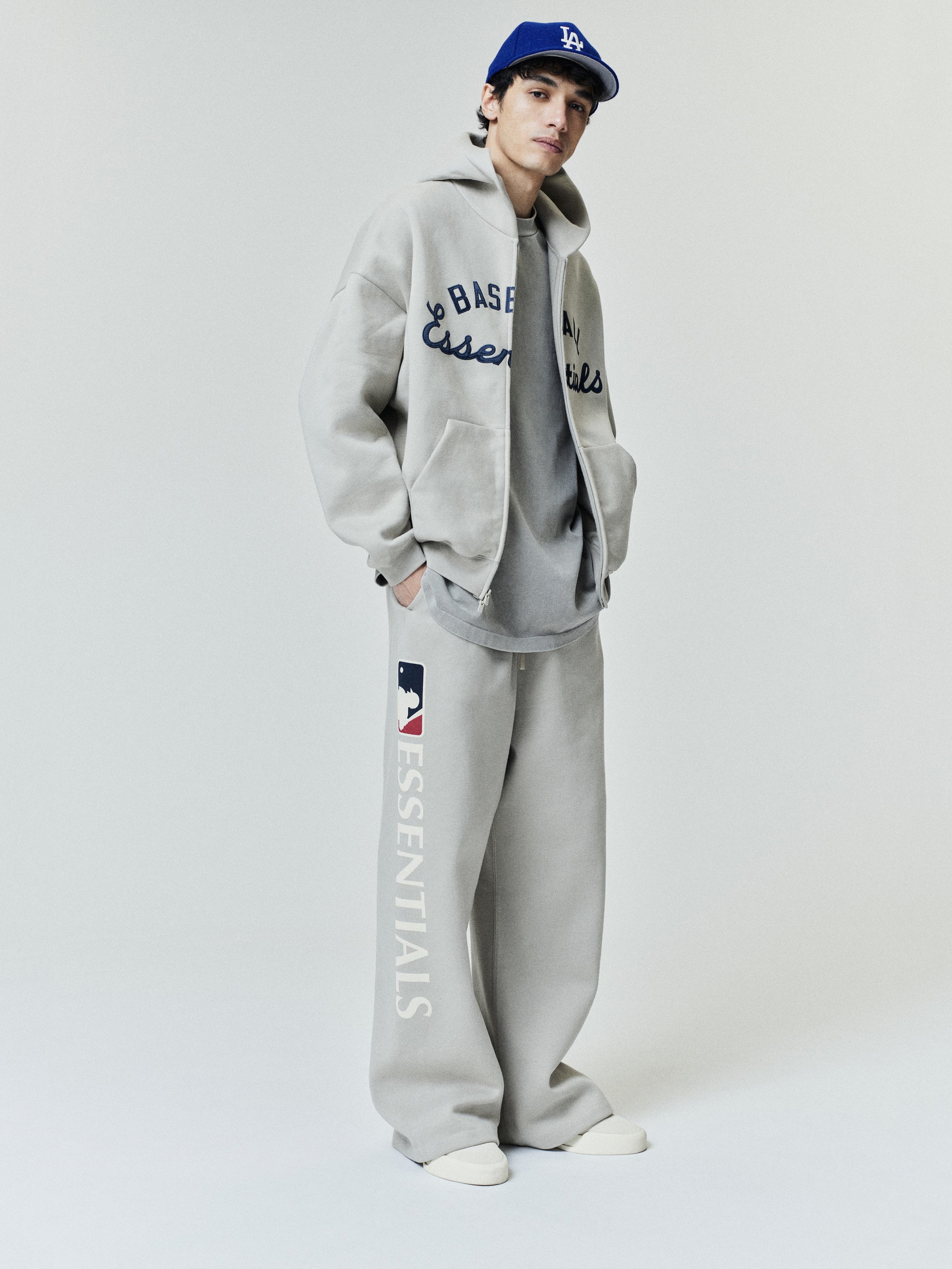 Model wearing Mlb Lounge Sweatpant in light gray with a relaxed fit and bold "ESSENTIALS" text and MLB logo on the side, paired with coordinating casual layered outerwear and sneakers.