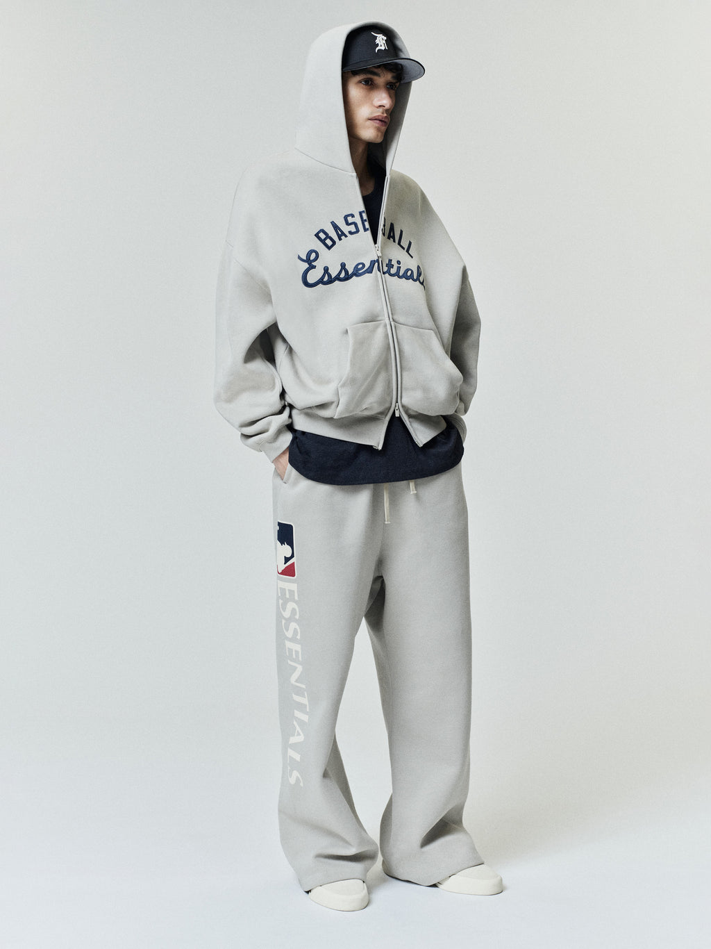 Model wearing Mlb Lounge Sweatpant in light gray with wide leg fit and large vertical "ESSENTIALS" text and MLB logo on the left leg, styled with matching zip-up hoodie and black baseball cap.