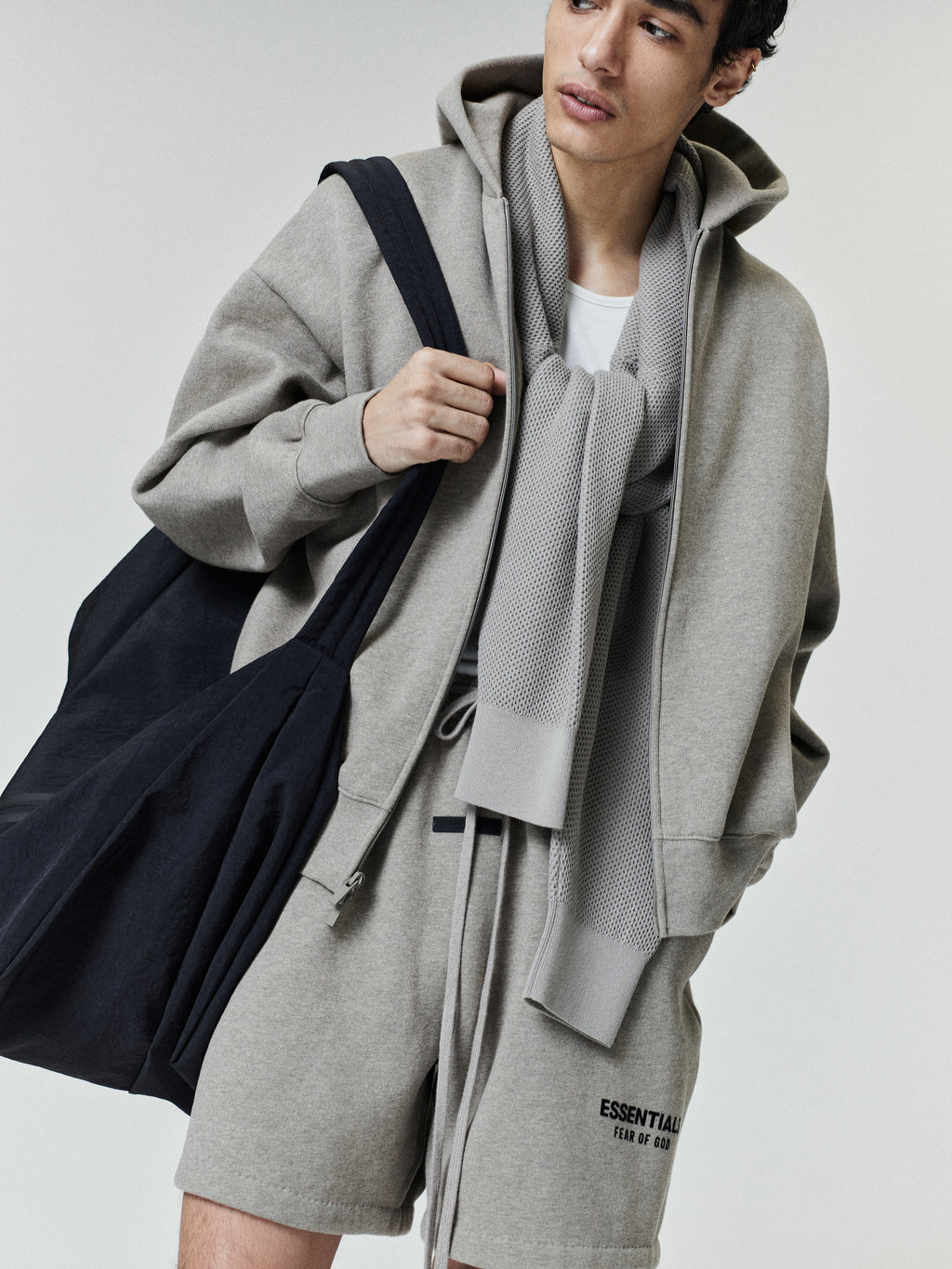 Young man wearing a light gray zip-up hoodie with matching shorts featuring small black text, a white t-shirt underneath, a textured gray scarf draped around the neck, and carrying a large black fabric shoulder bag.