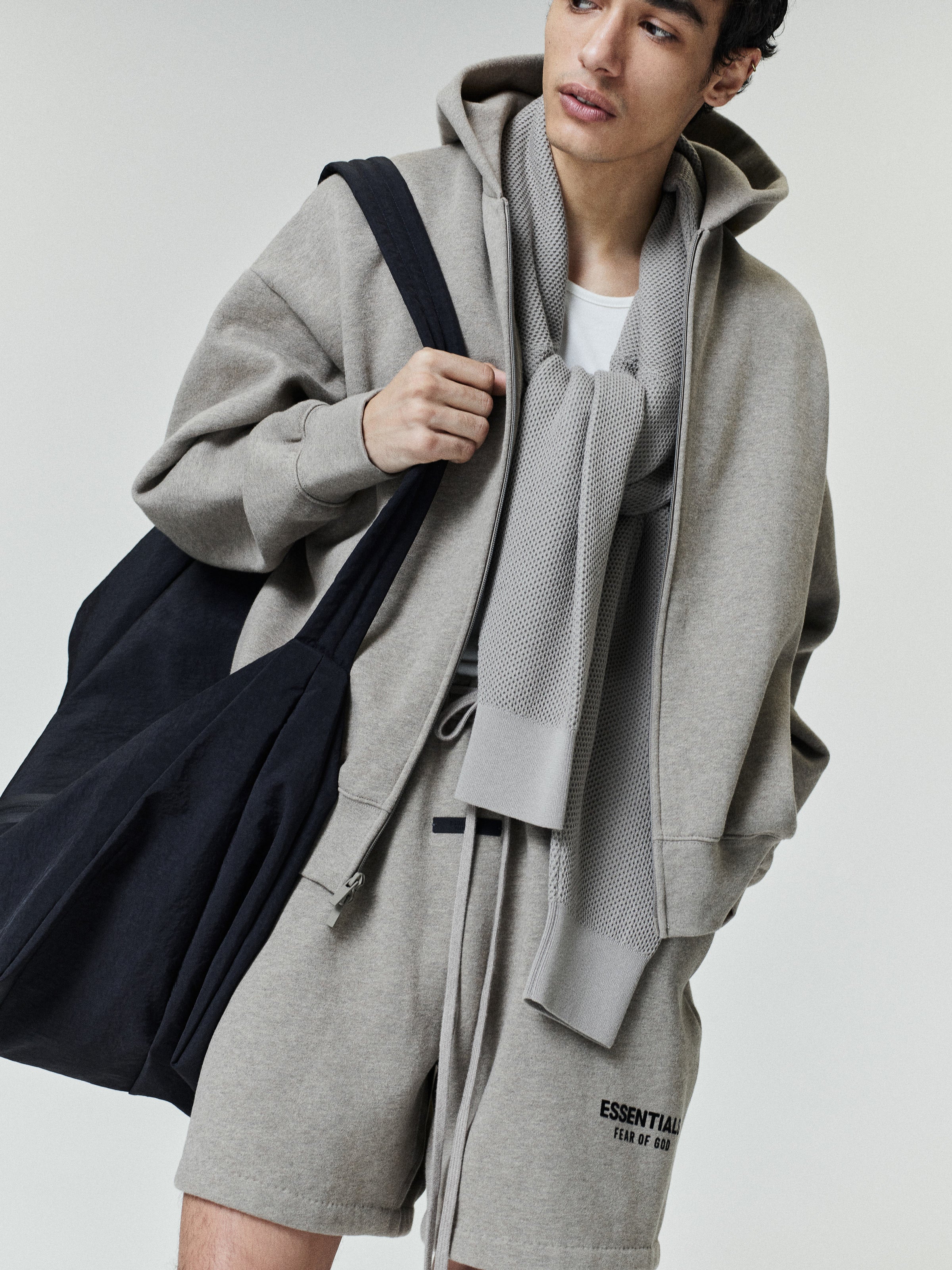 Young man wearing a light gray zip-up hoodie with matching drawstring shorts featuring small black text, a white t-shirt underneath, a textured gray scarf around the neck, and carrying a large black fabric tote bag.