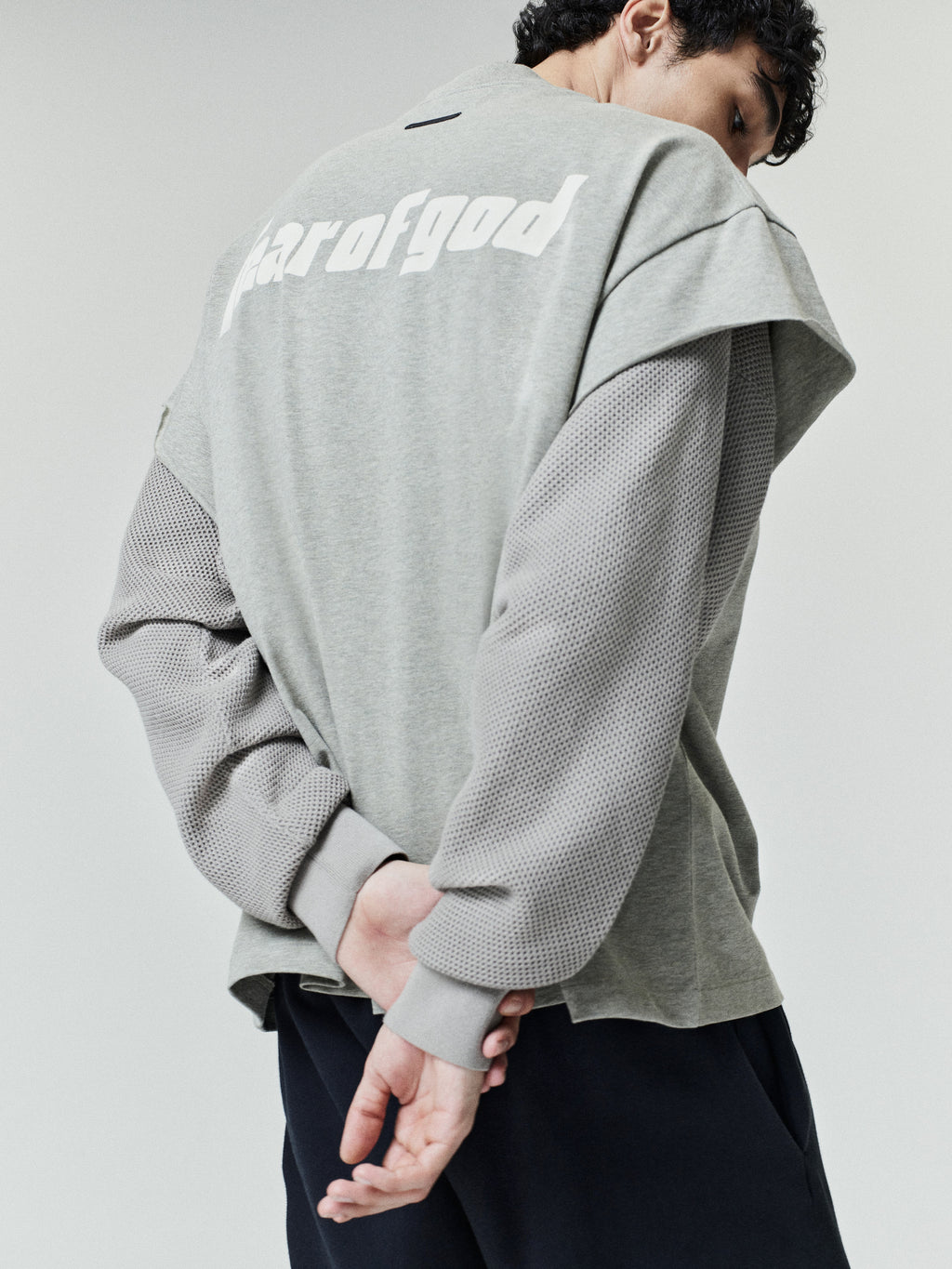 Rear view of a model wearing the Fear Of God X Franklin 90s Cutoff with a soft gray body and textured mesh long sleeves, paired with relaxed dark pants.