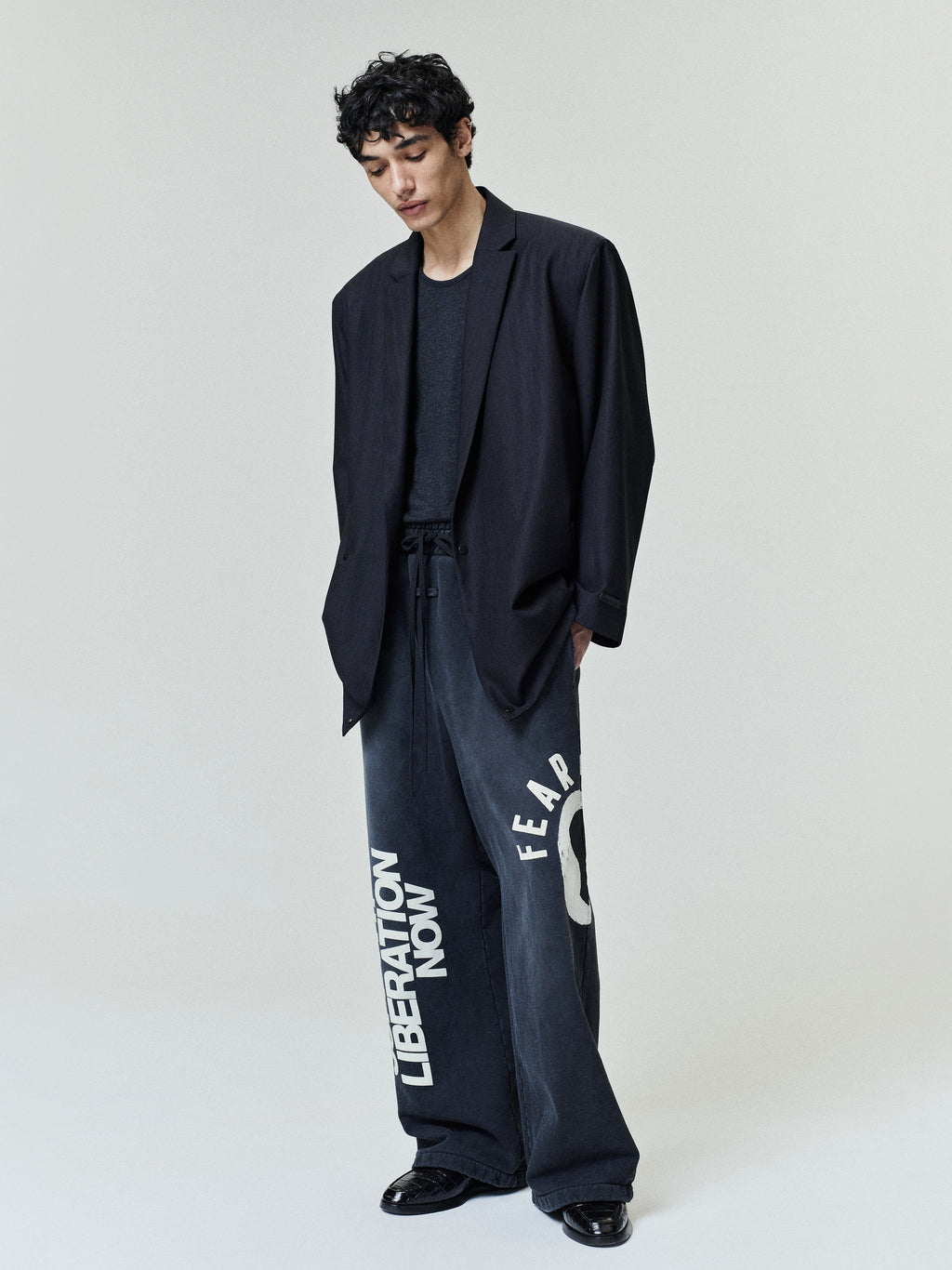 Young man wearing a loose black blazer over a dark t-shirt paired with oversized dark pants featuring bold white graphic text on the legs, standing against a plain light background.