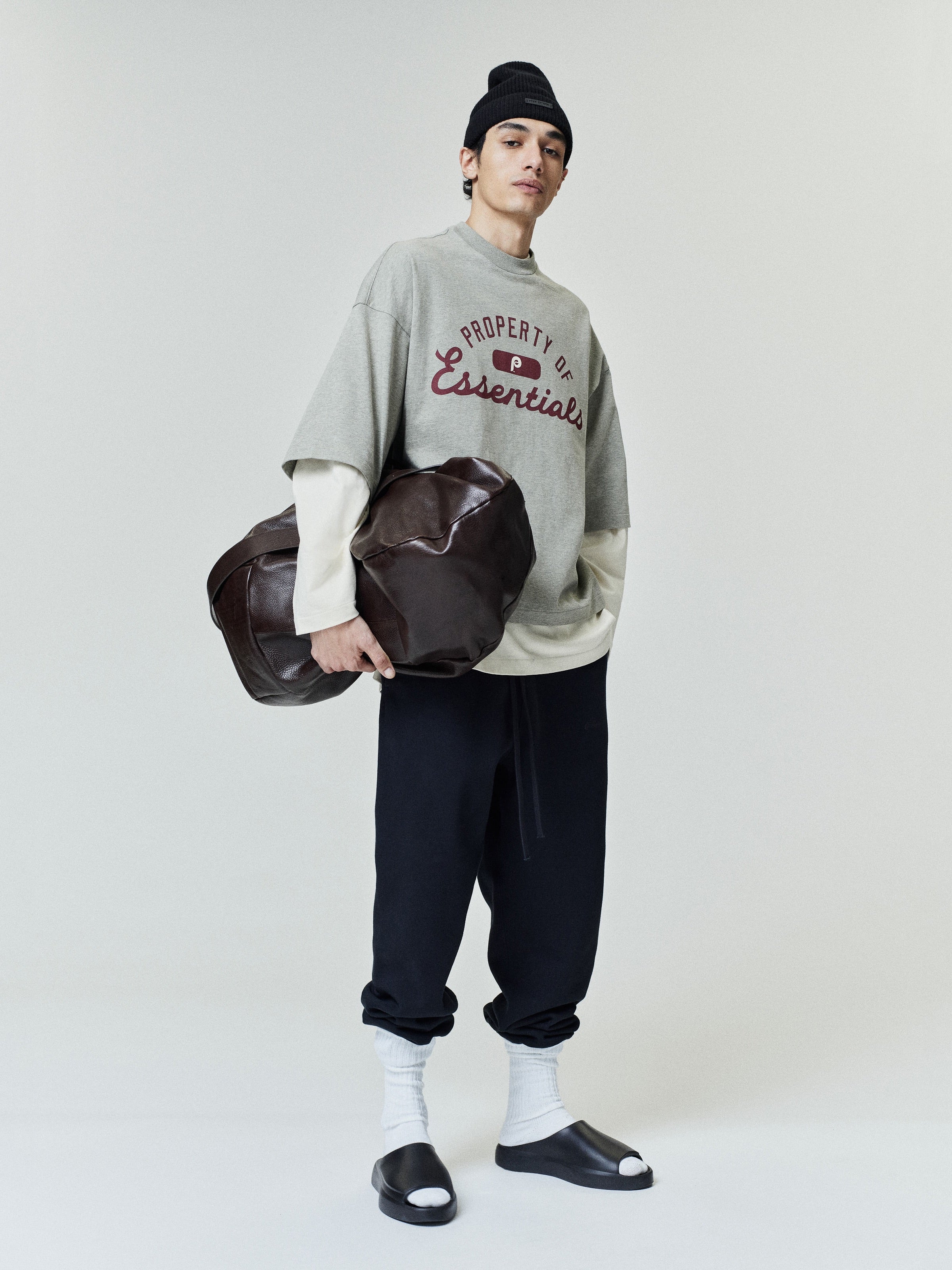 Model wearing a loose-fit Phillies Merch Tee in muted gray layered over a long-sleeve cream shirt, paired with black relaxed pants, white socks, black slide sandals, and a black beanie, holding a large brown leather duffel bag.