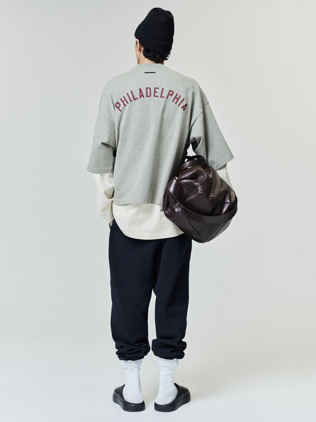 Model seen from behind wearing the Phillies Merch Tee in light gray with "PHILADELPHIA" text on the back, layered over a cream shirt, paired with dark relaxed pants, white socks, black slides, a black beanie, and carrying a large brown leather bag.