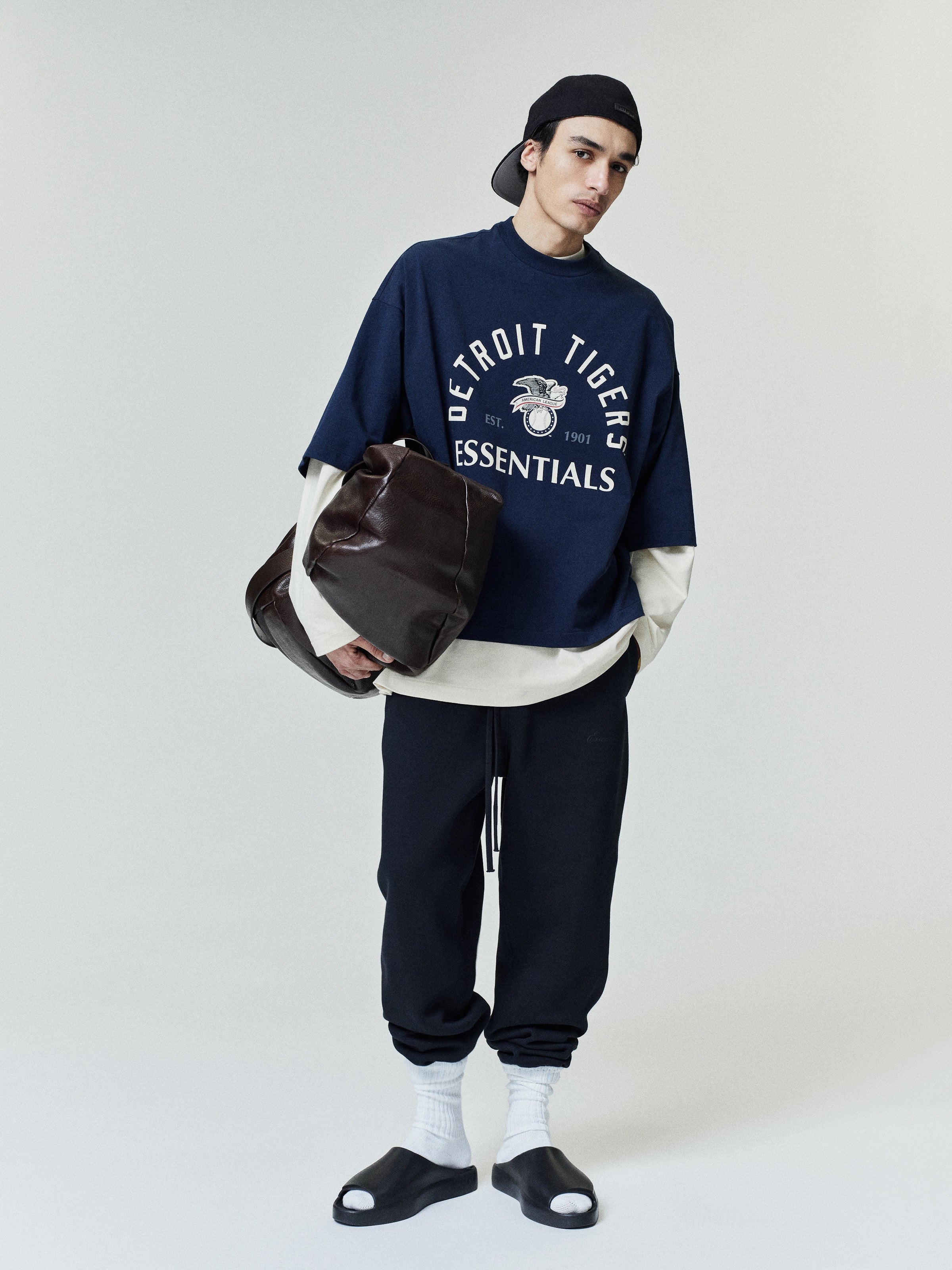 Person wearing a navy 'Detroit Tigers Essentials' sweatshirt with a white shirt underneath, dark pants, and black shoes on a light gray background.