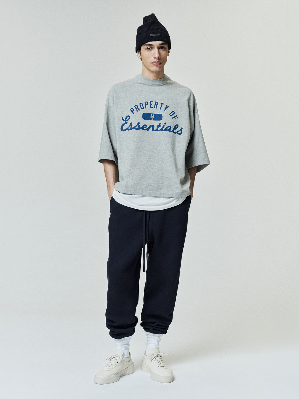 Model wearing a grey Mets Merch Tee with "Property of Essentials" printed in blue on the front, styled with layered tops, black joggers, white socks, white sneakers, and a black beanie.