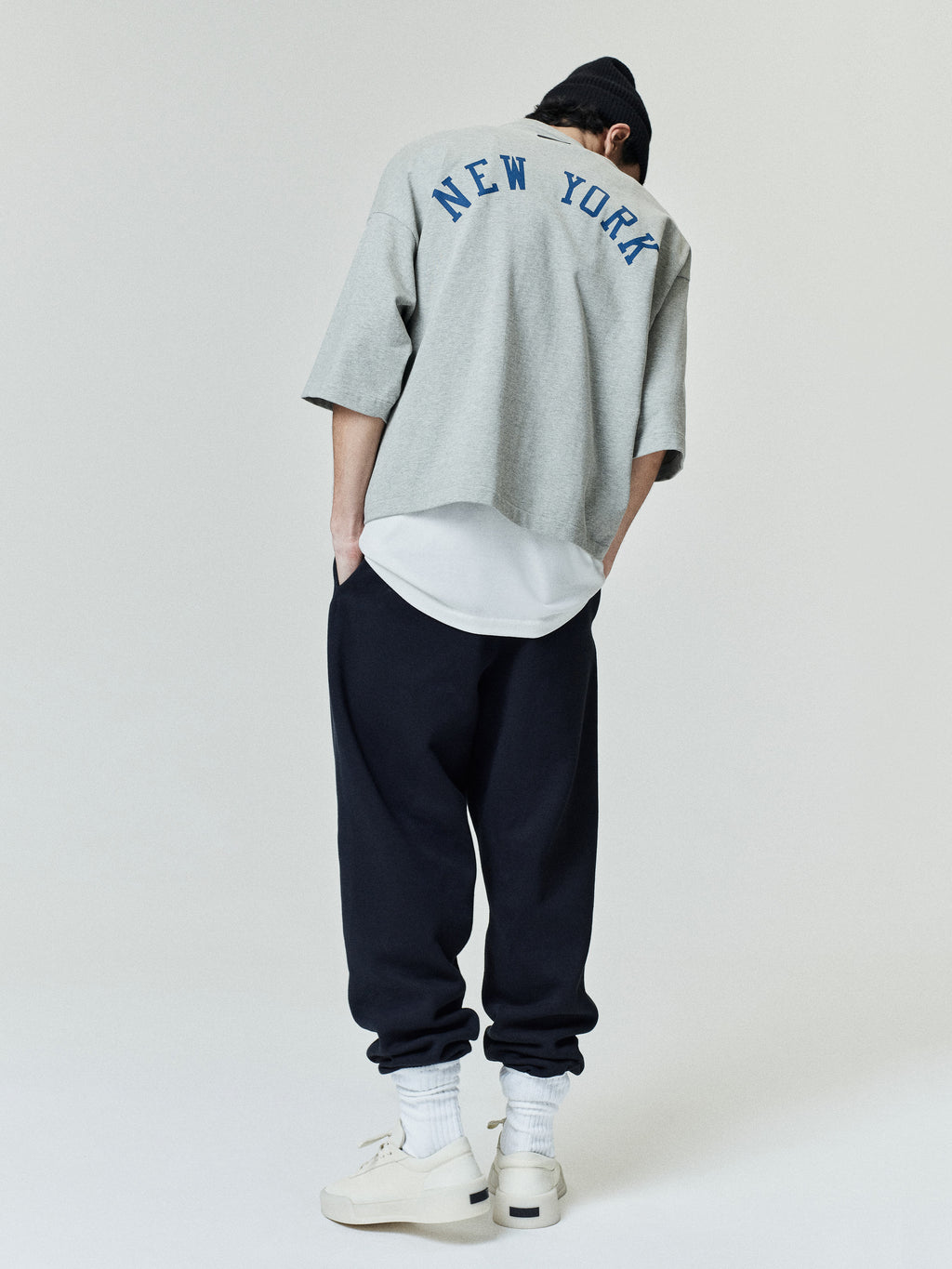 Rear view of a model wearing a grey Mets Merch Tee with "NEW YORK" printed in blue across the upper back, styled with dark pants, white sneakers, and a black beanie in a casual look.