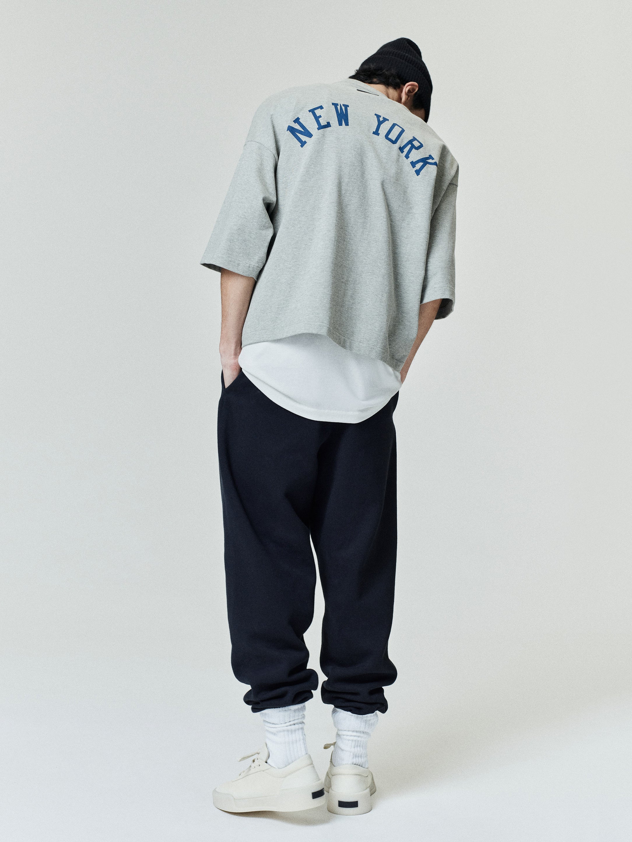 Rear view of a model wearing a grey Mets Merch Tee with "NEW YORK" printed in blue across the upper back, styled with dark pants, white sneakers, and a black beanie in a casual look.
