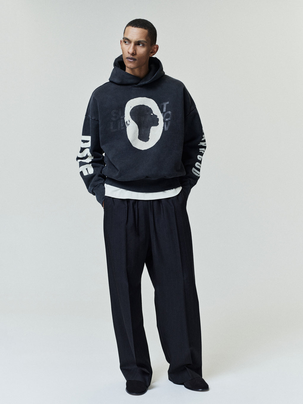 Man wearing a black hoodie with a large white silhouette profile graphic on the front and white text printed on both sleeves, paired with loose black trousers and a white shirt layered underneath against a plain background.