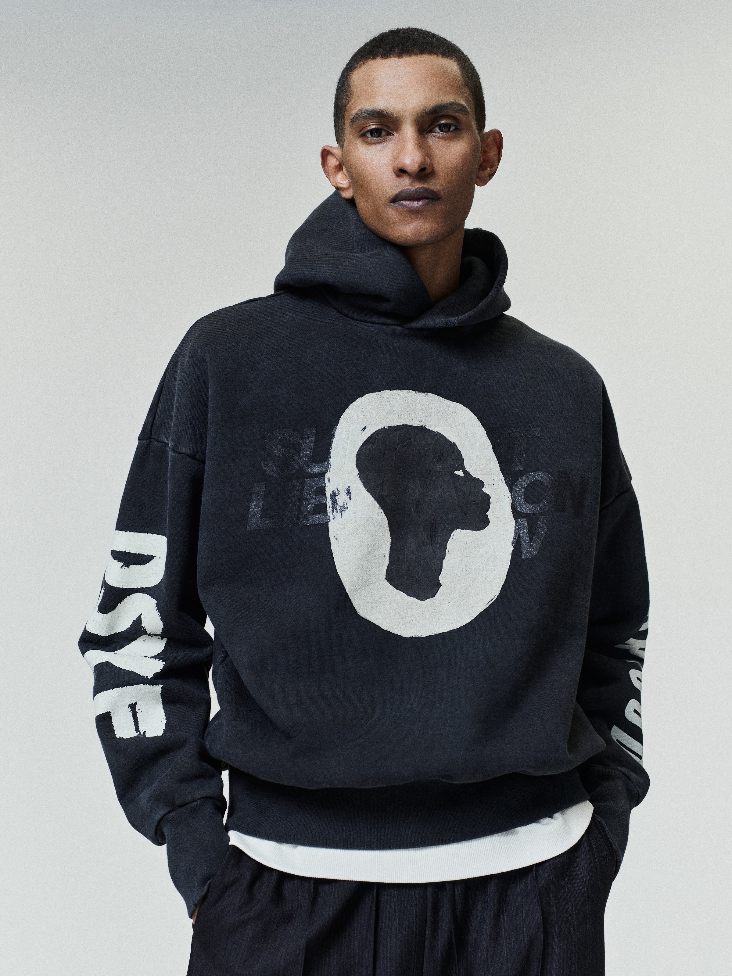 Man wearing a faded black hoodie featuring a large white profile silhouette graphic on the front and bold white text printed along the sleeves, paired with black pants and a layered white shirt underneath against a plain background.