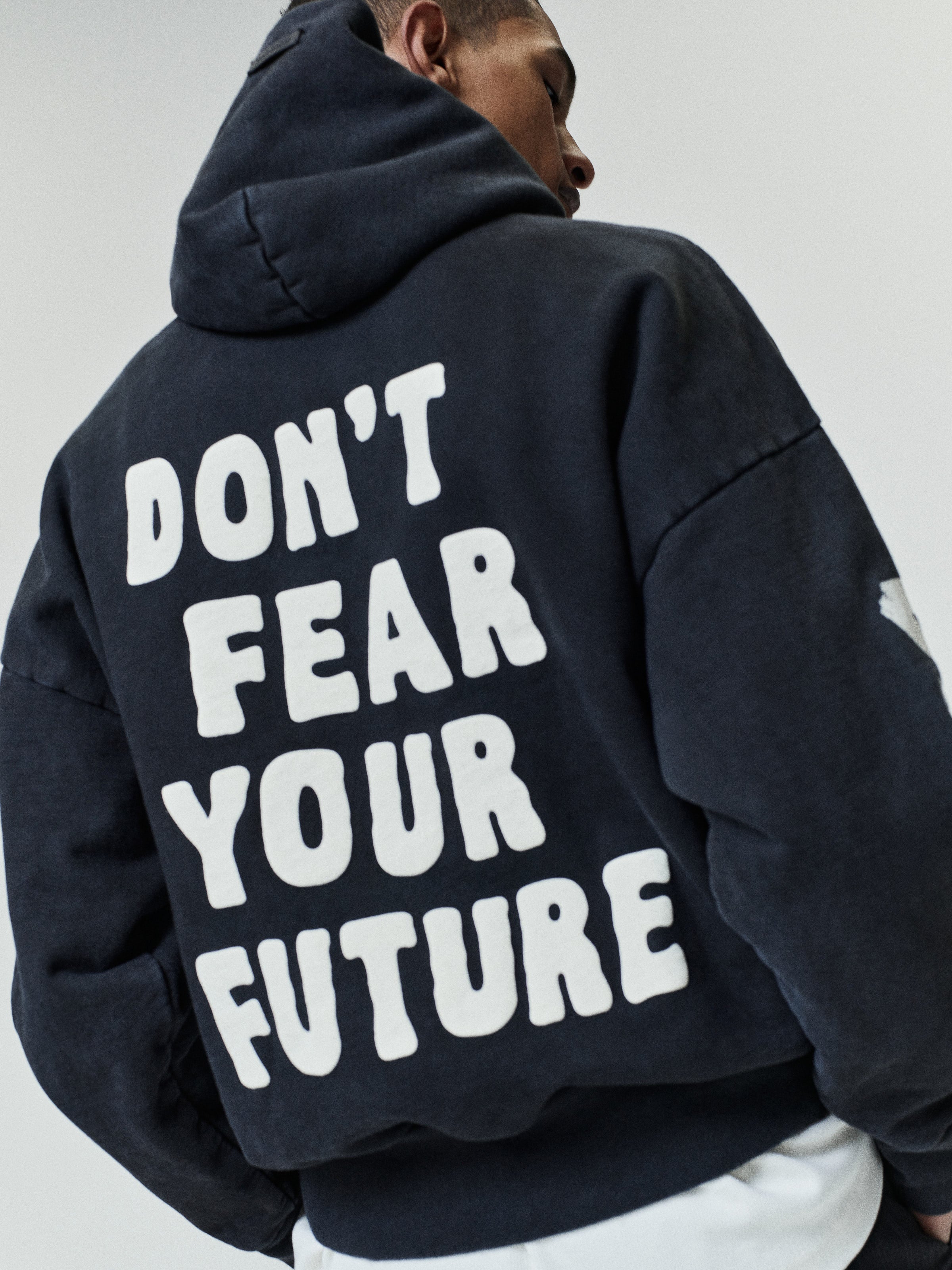 Person wearing a dark oversized hoodie with bold white text printed on the back that reads "DON'T FEAR YOUR FUTURE," shown from behind against a neutral background