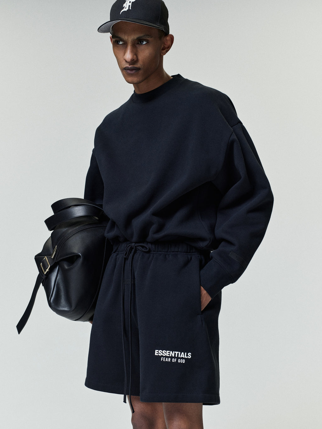 Slim man wearing a loose black sweatshirt and matching shorts with white "ESSENTIALS FEAR OF GOD" text on the left leg, a black cap, and holding a large black leather bag.