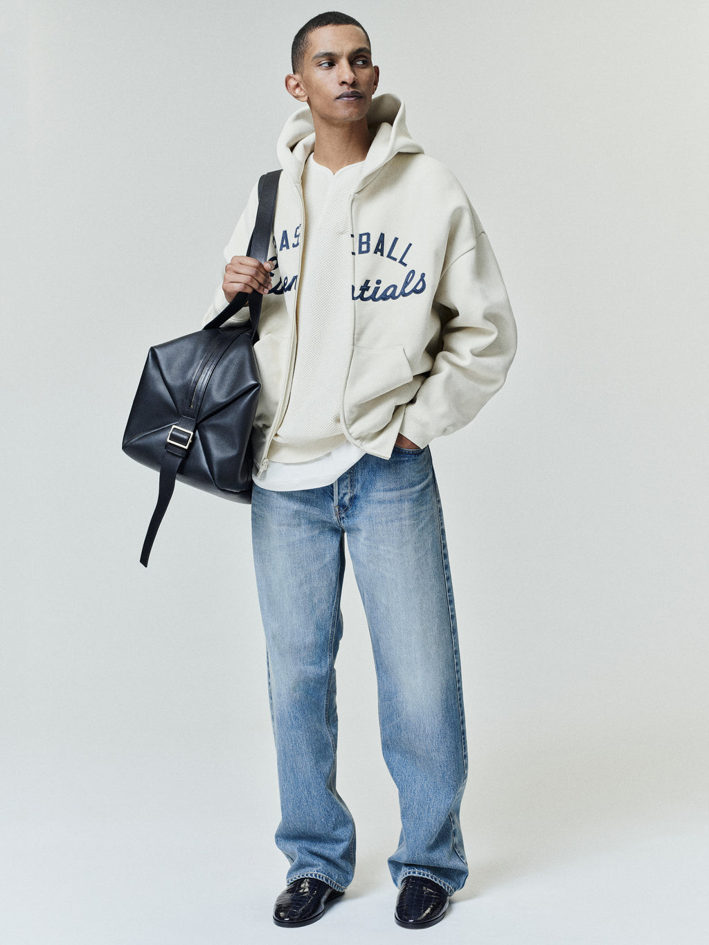 Model wearing an off-white Mlb Full Zip Sport Hoodie with bold navy lettering across the chest, paired with light-wash jeans and black leather shoes, holding a black leather bag.