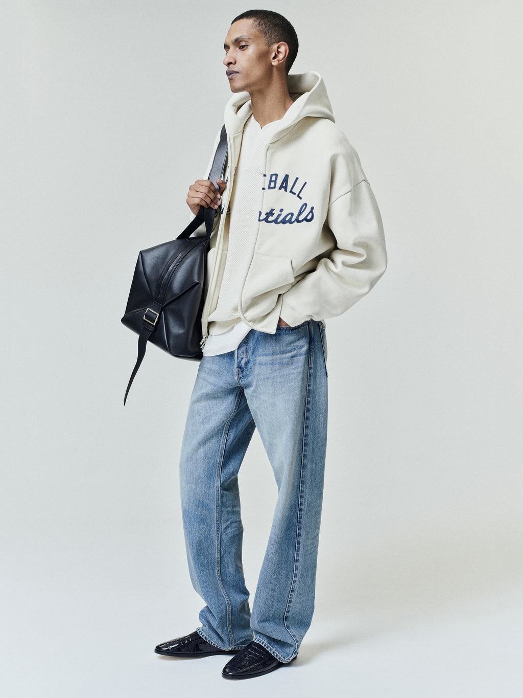 Model wearing an off-white Mlb Full Zip Sport Hoodie with navy script detailing, paired with light blue jeans and black leather shoes, accessorized with a black leather shoulder bag.